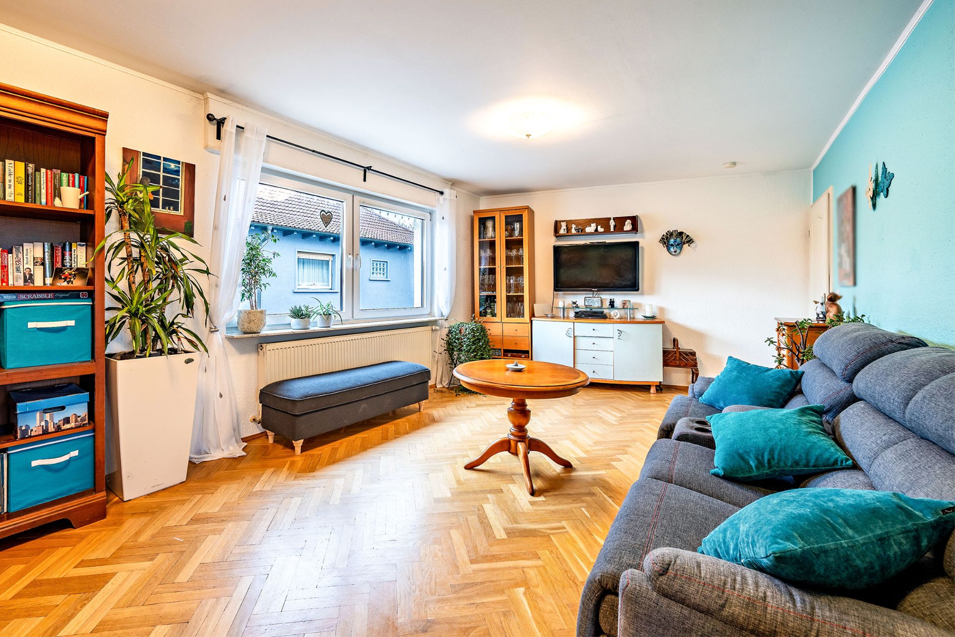 Helles Wohnzimmer mit grauem Sofa, Holztisch, Bücherregal, Fenster und Fernseher.
