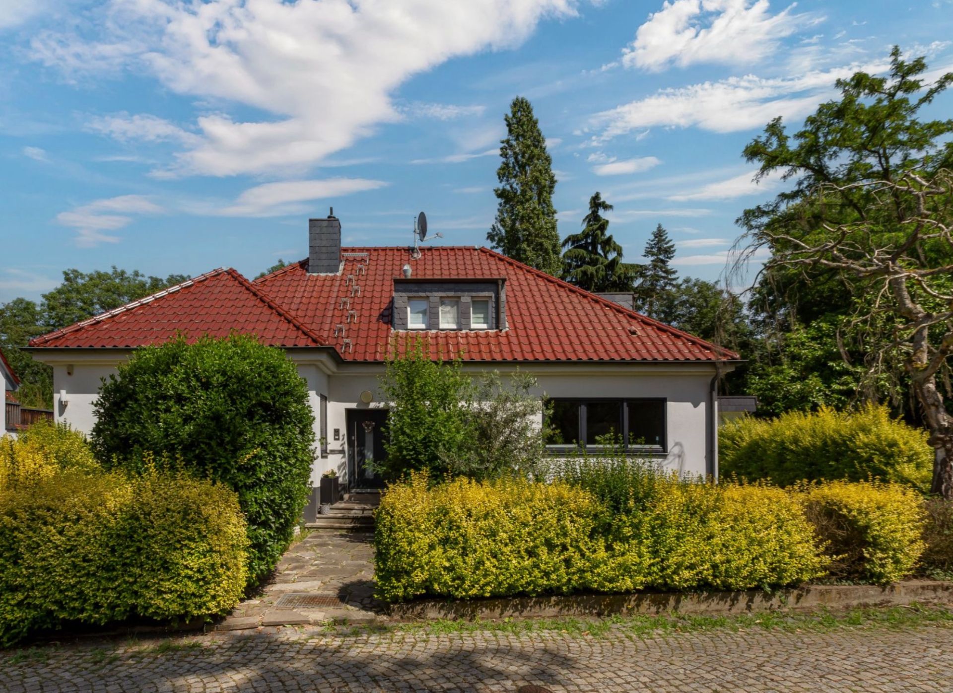 Einfamilienhaus mit rotem Dach, großen Fenstern und Garten mit Büschen, Gehweg führt zum Eingang.