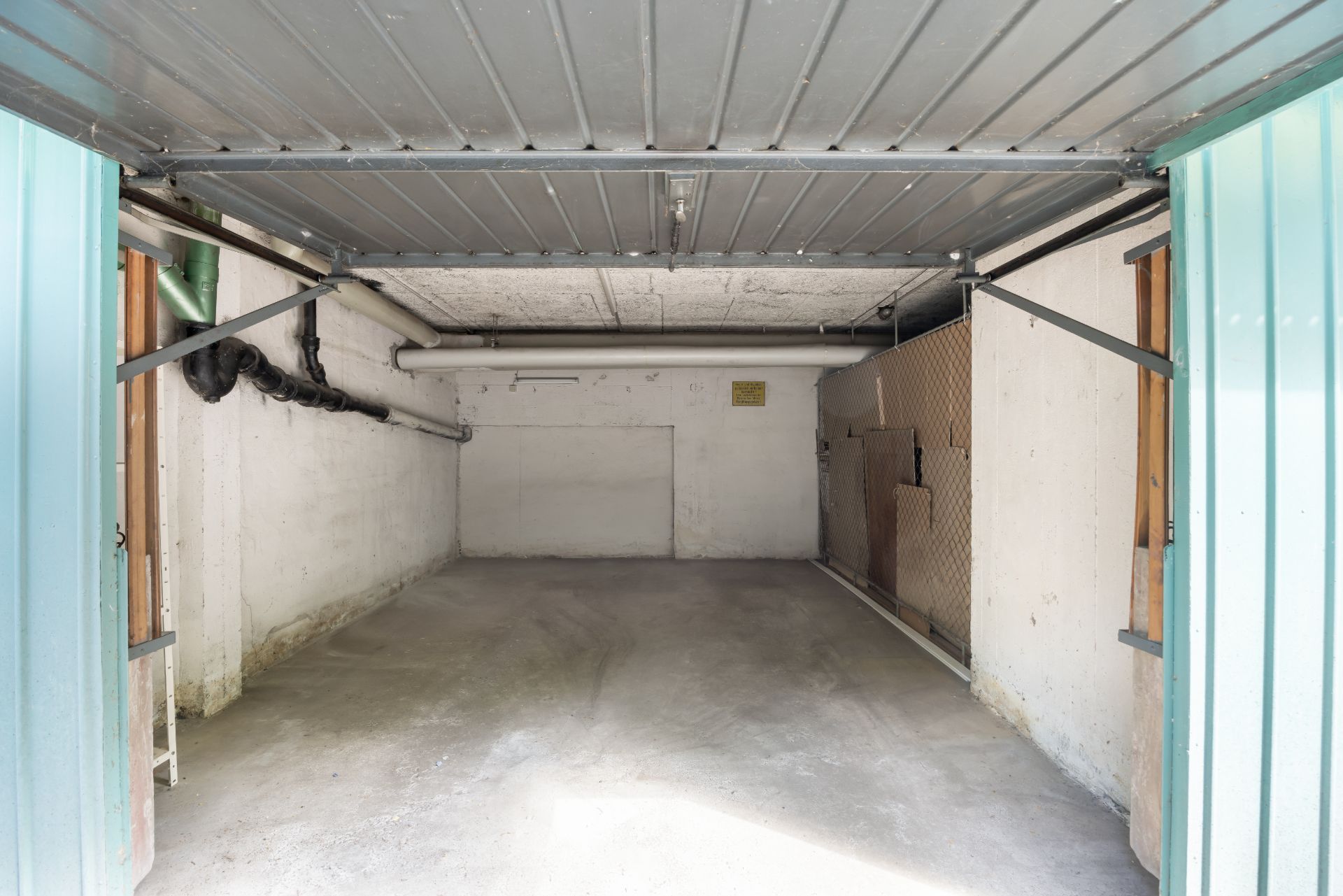 Leere Garage mit geöffnetem Tor, Betonboden und sichtbaren Rohren an der Decke.