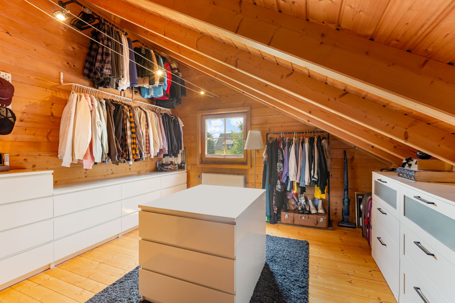 Helles Ankleidezimmer mit Holzdecke, Kleiderschränken, Kommoden und einem Fenster.