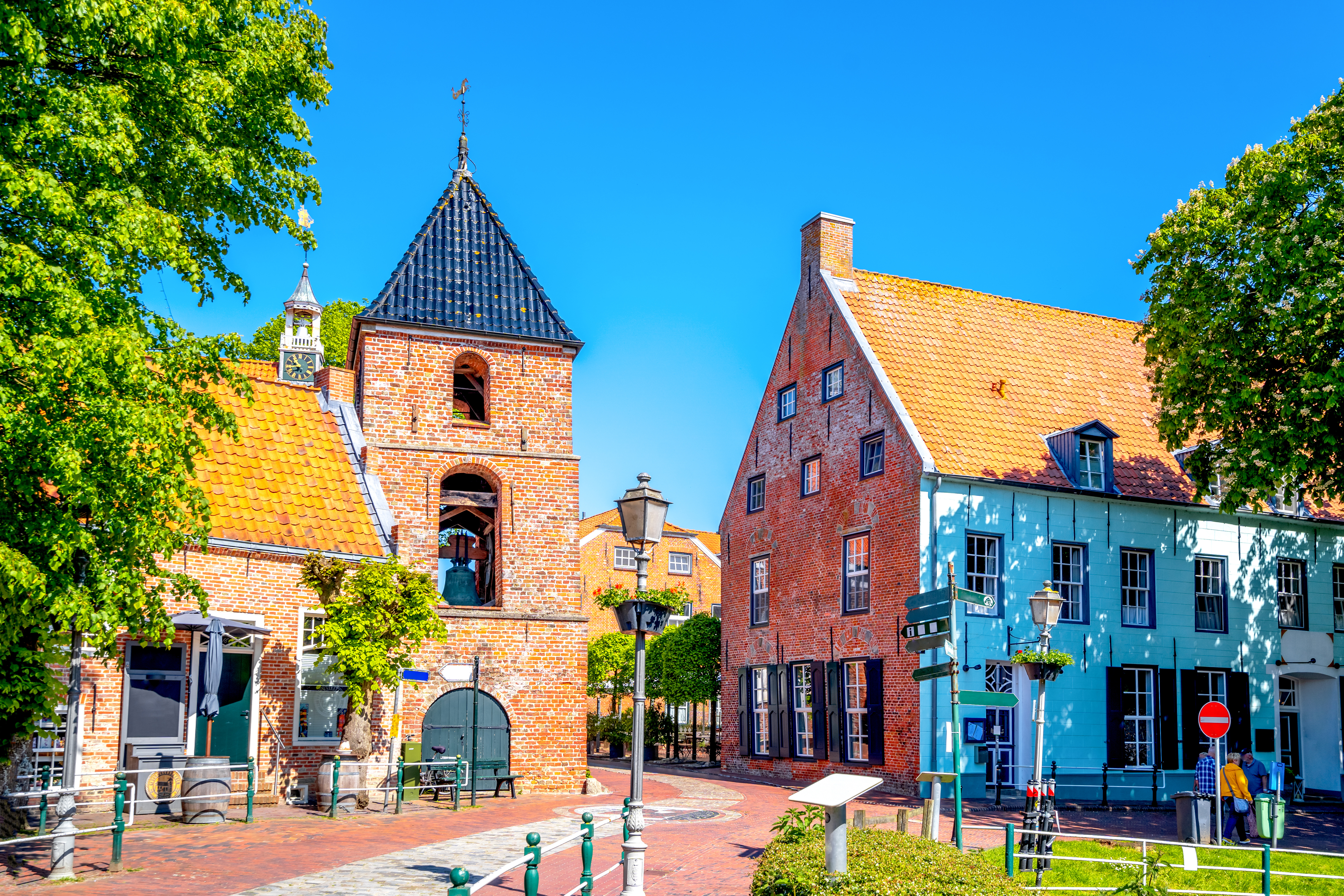 Historische Backsteinhäuser mit roten Dächern an einem sonnigen Platz, Bäume und Straßenlaternen sichtbar.