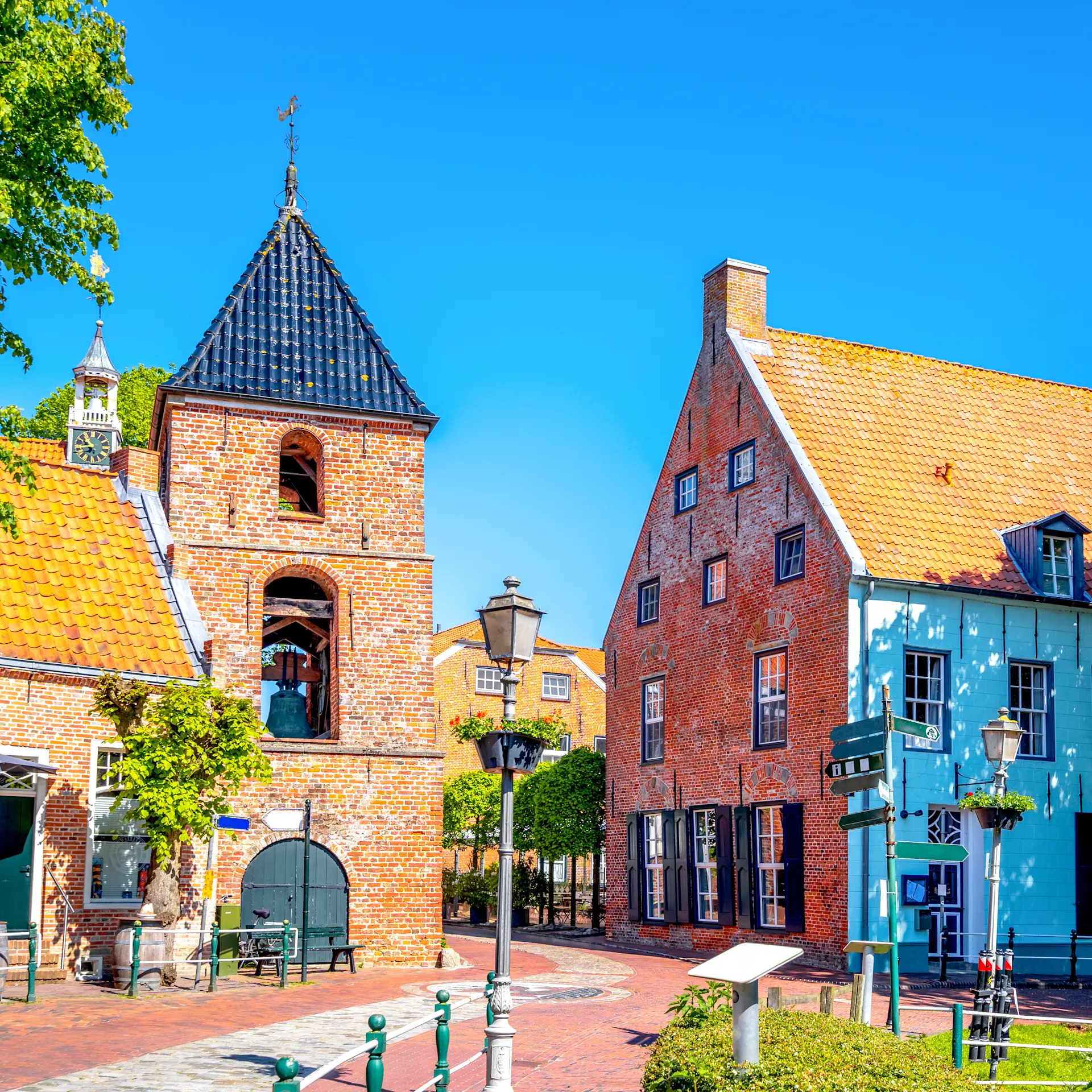 Historische Backsteinhäuser mit roten Dächern an einem sonnigen Platz, Bäume und Straßenlaternen sichtbar.