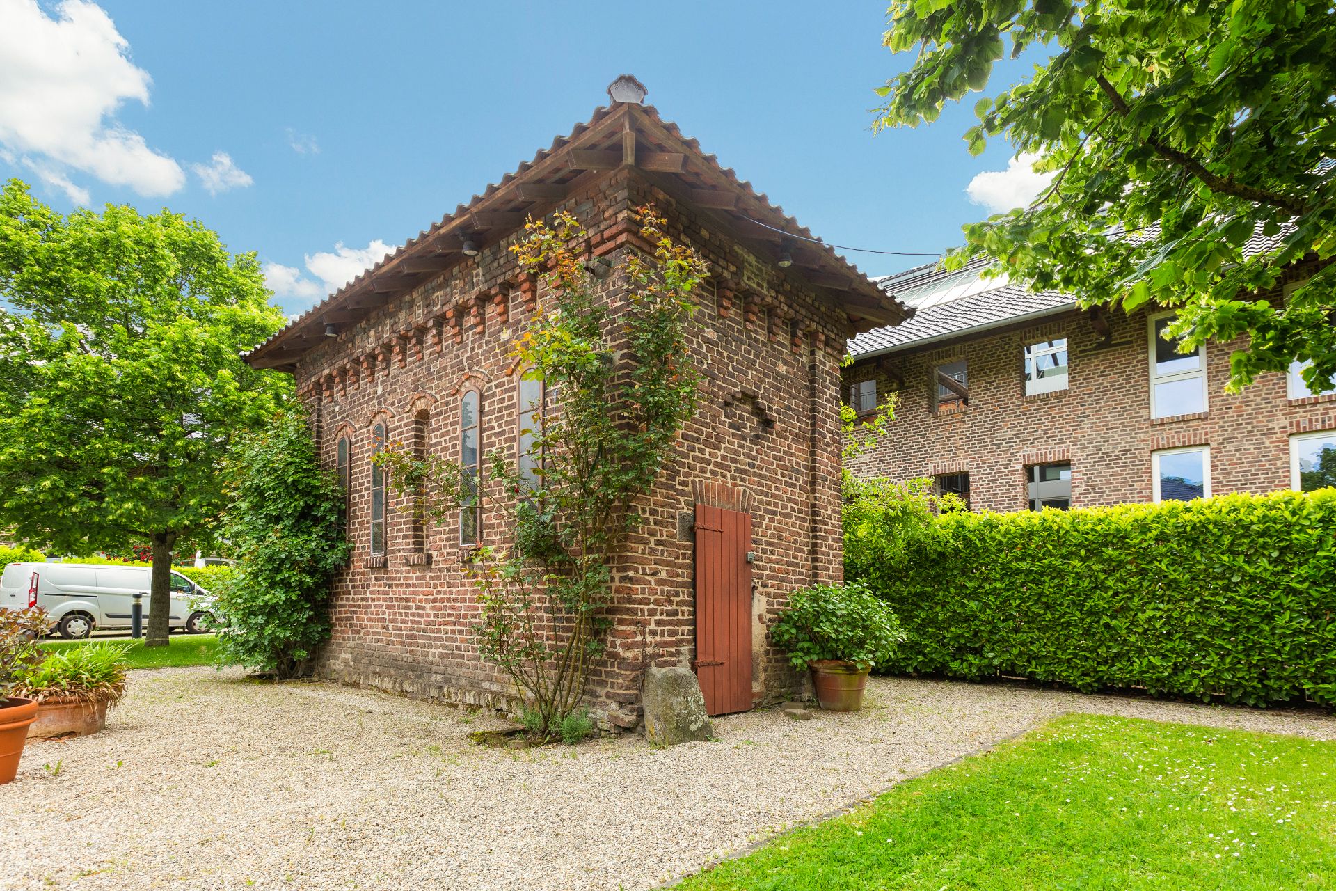 Kleines Backsteingebäude im Garten mit Holztür und Fenstern, umgeben von Büschen und Bäumen.