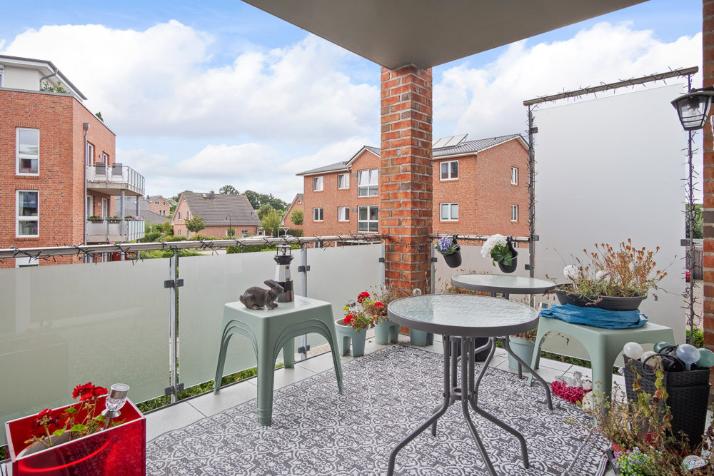 Überdachter Balkon mit kleinem Tisch, Stühlen, Blumen und Blick auf rote Wohngebäude.