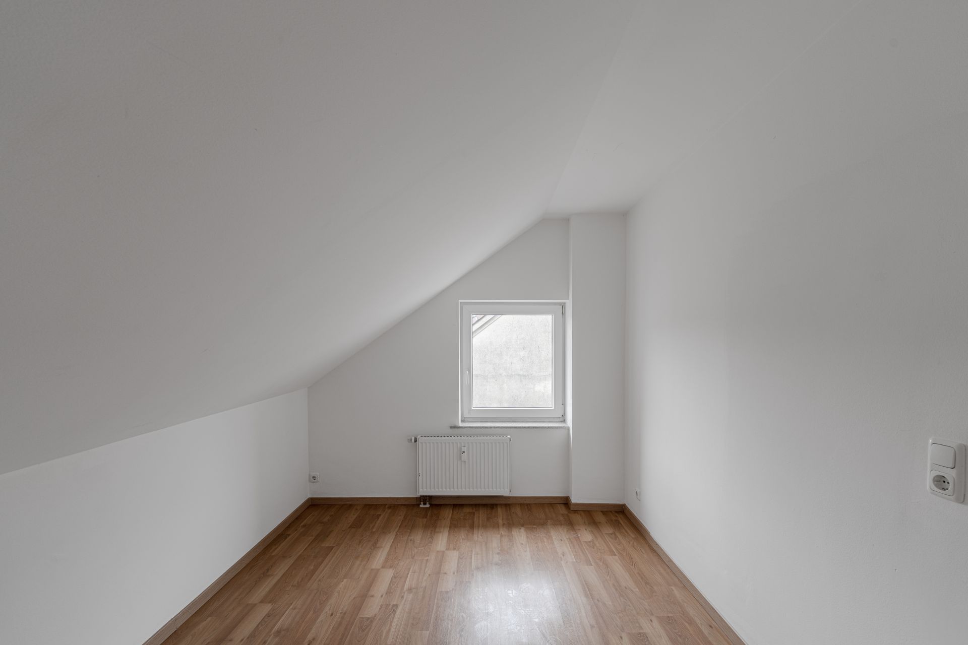 Leerer Raum mit Dachschräge, weiß gestrichen, Holzfußboden, Fenster und Heizkörper.