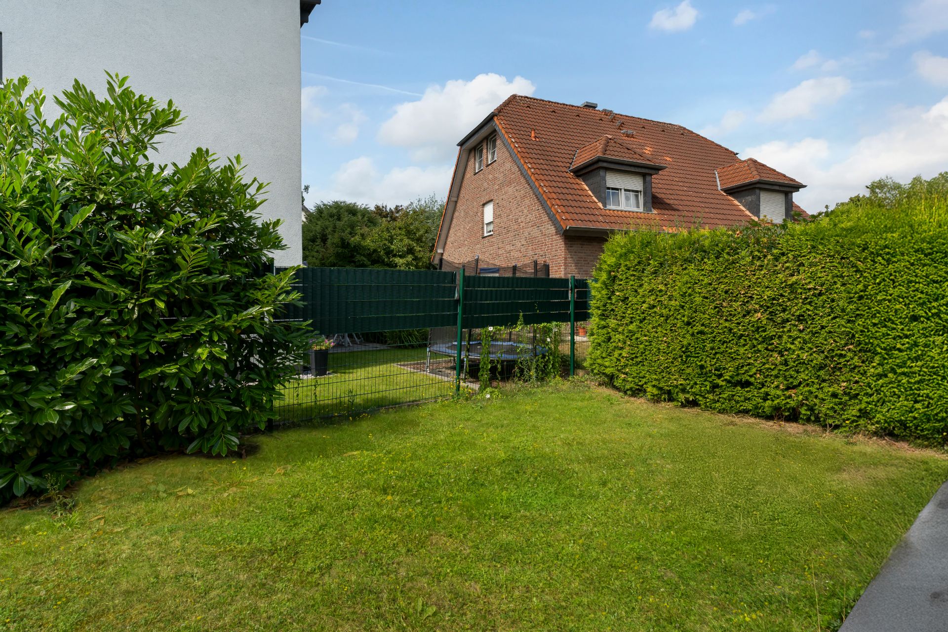 Garten mit Rasenfläche, grüner Hecke und Blick auf ein Wohnhaus mit rotem Dach.