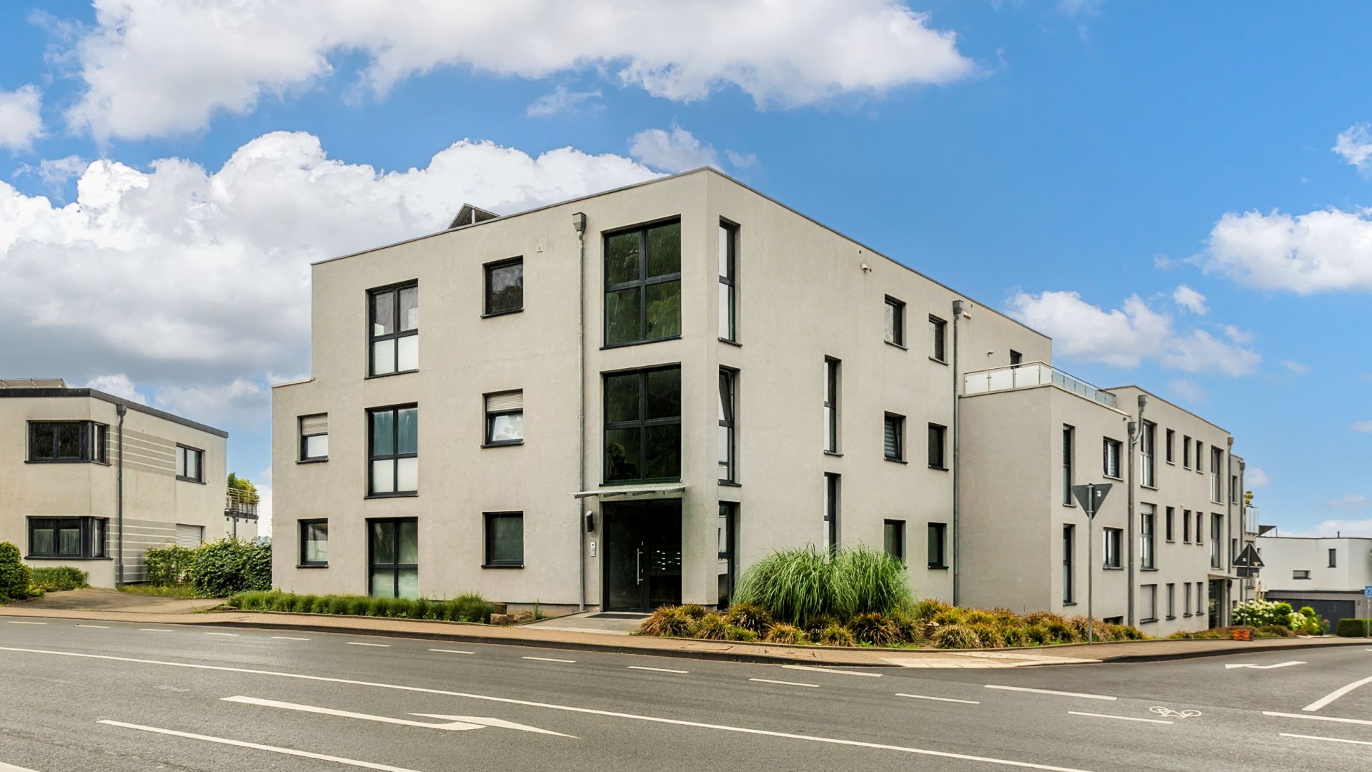Modernes, weißes Wohngebäude mit großen Fenstern an einer Straßenecke, umgeben von Pflanzen.