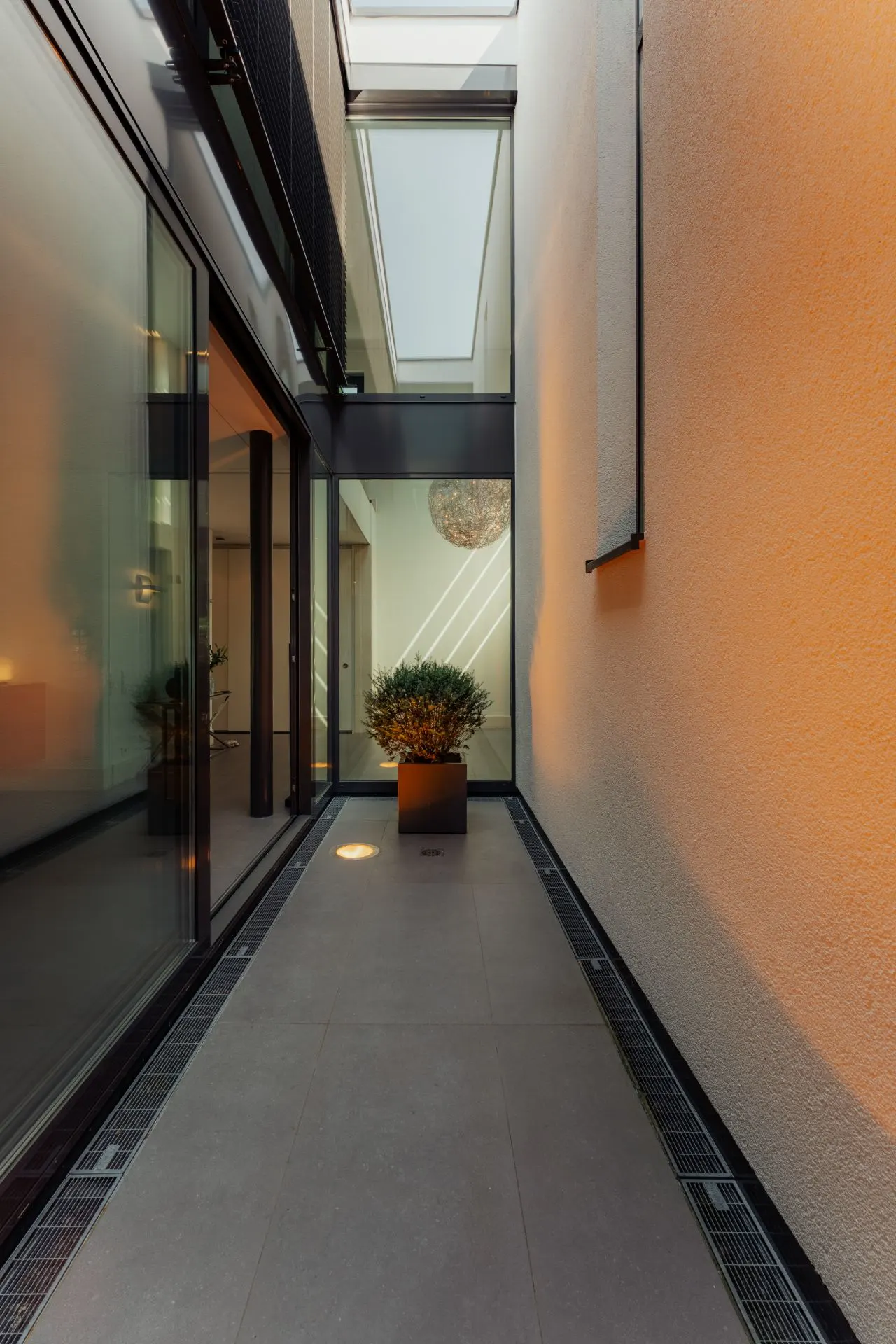 Moderner, schmaler Hausflur mit Glaswänden, Oberlichtern und einem Pflanzkübel am Ende.