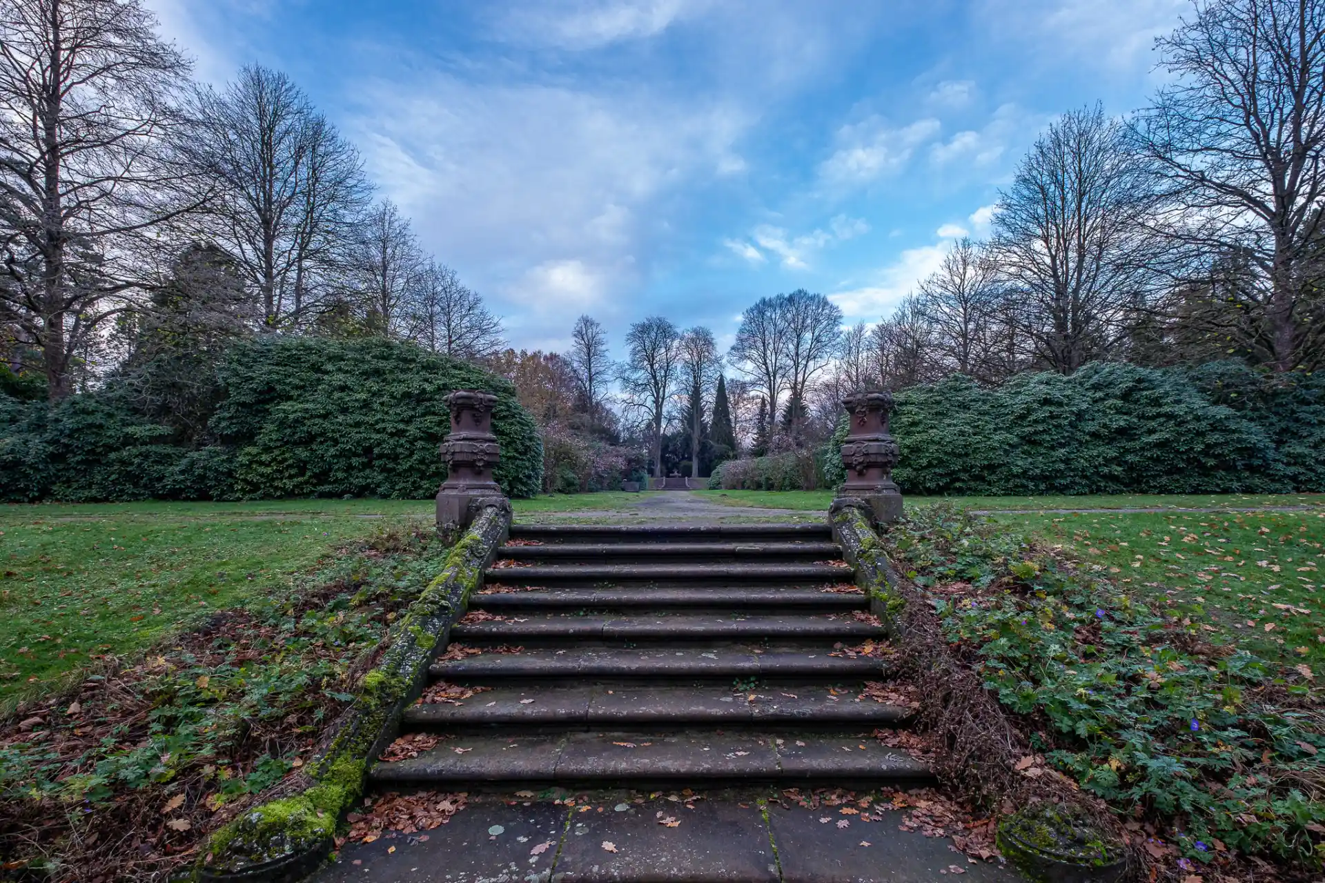 Steintreppe im Garten mit Moos und Laub, flankiert von zwei steinernen Vasen, umgeben von Bäumen.