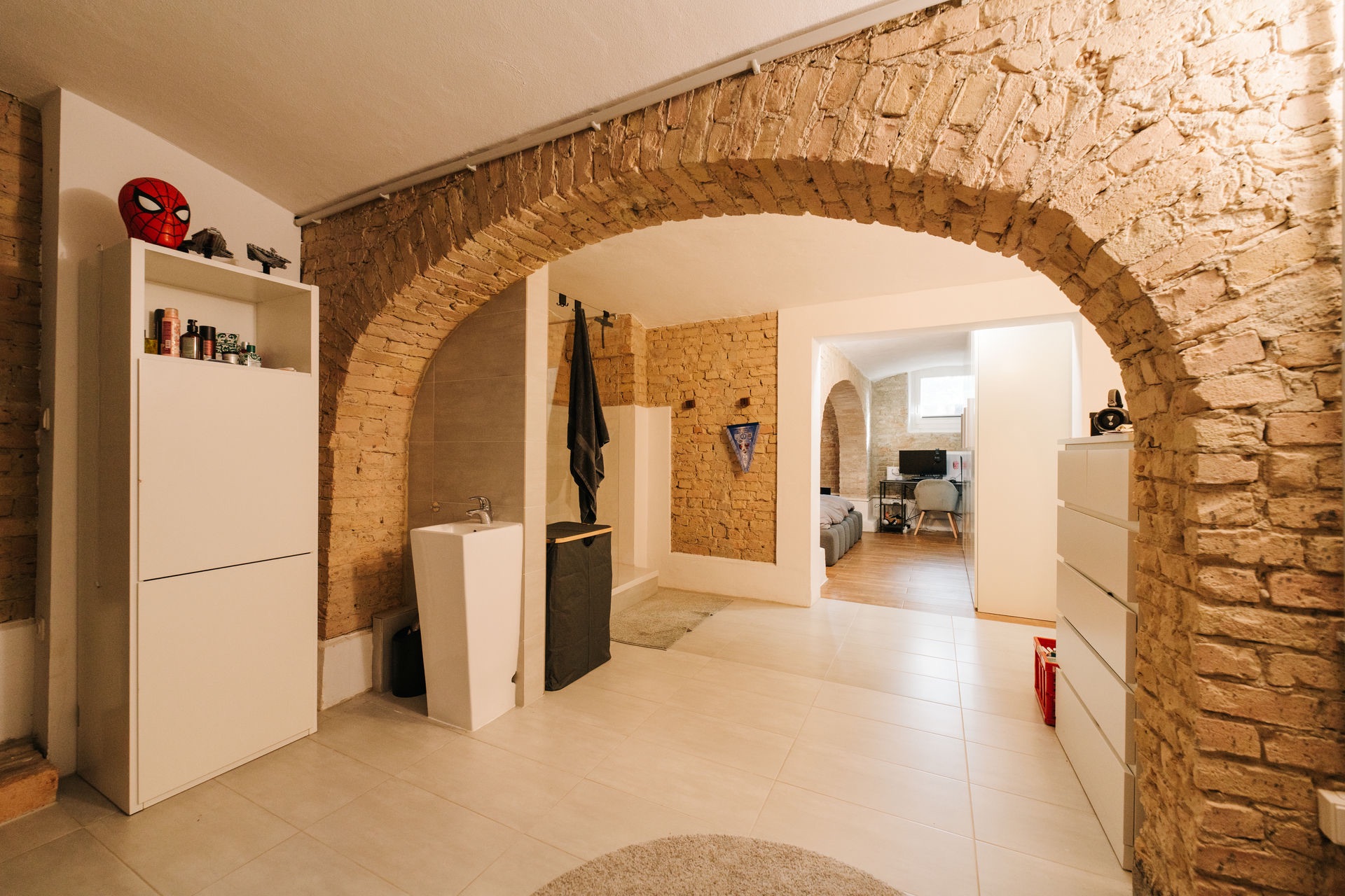 Raum mit Backsteinbogen, weißen Schränken, Waschbecken und Blick in ein Schlafzimmer mit Schreibtisch.