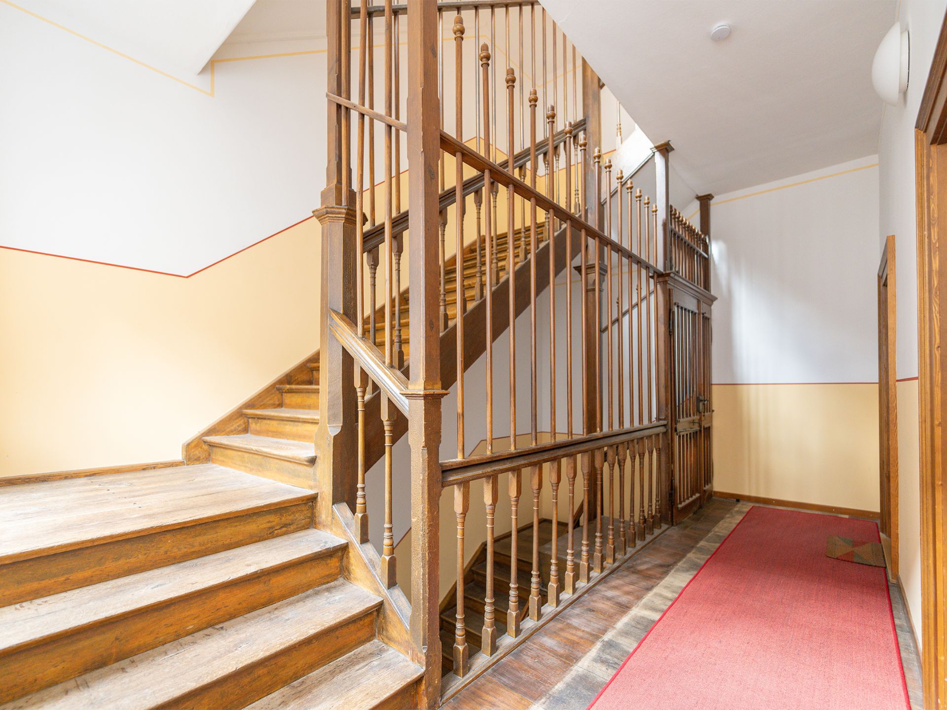 Helles Treppenhaus mit Holzgeländer und rotem Teppichläufer im Flur.