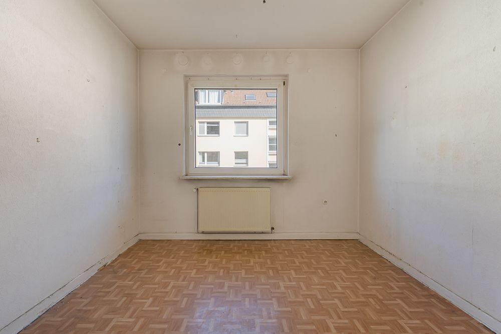 Leeres Zimmer mit Parkettboden, weiß gestrichenen Wänden und einem Fenster mit Heizung darunter.