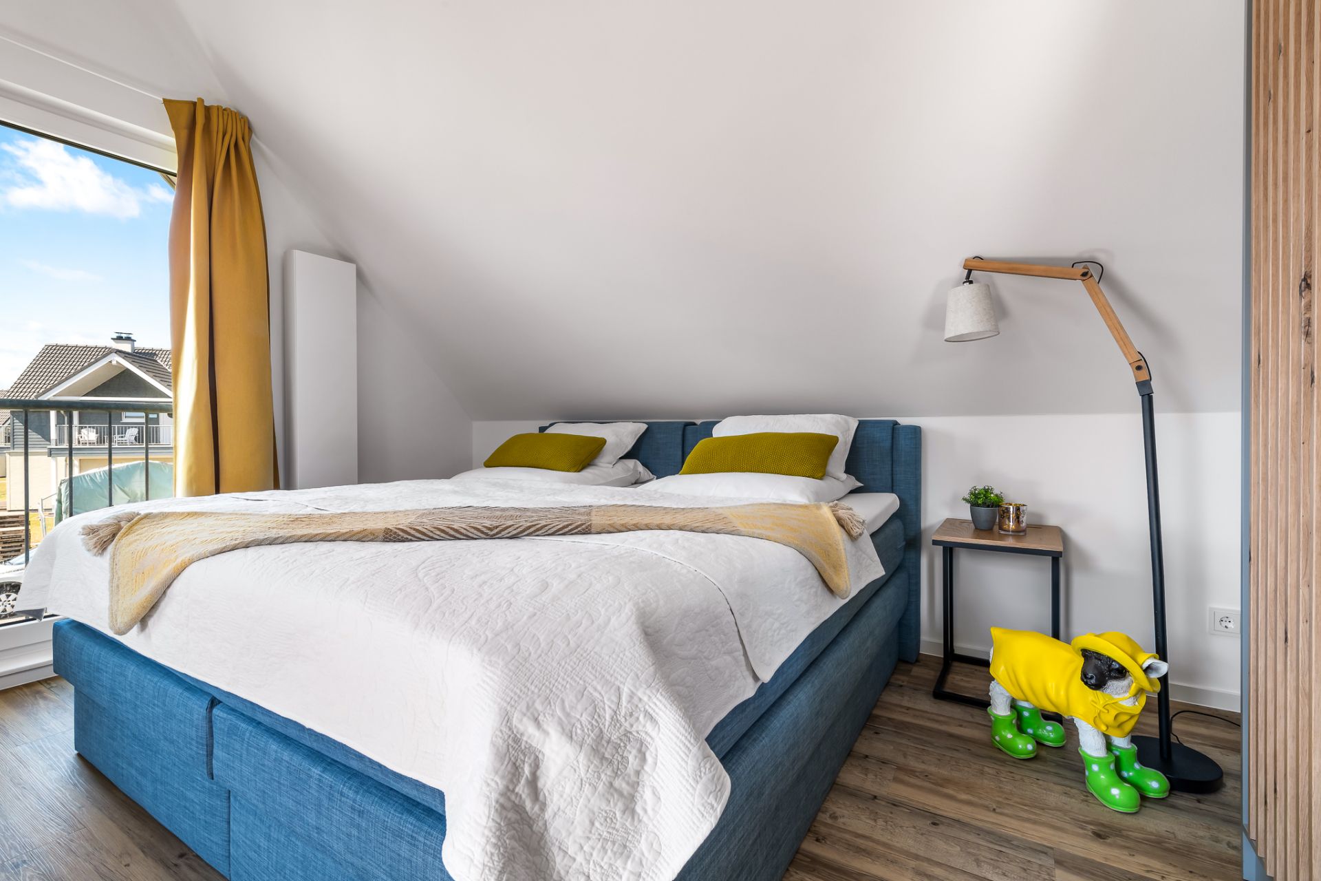 Schlafzimmer mit blauem Bett, gelben Kissen, Nachttisch und Stehlampe unter Dachschräge.