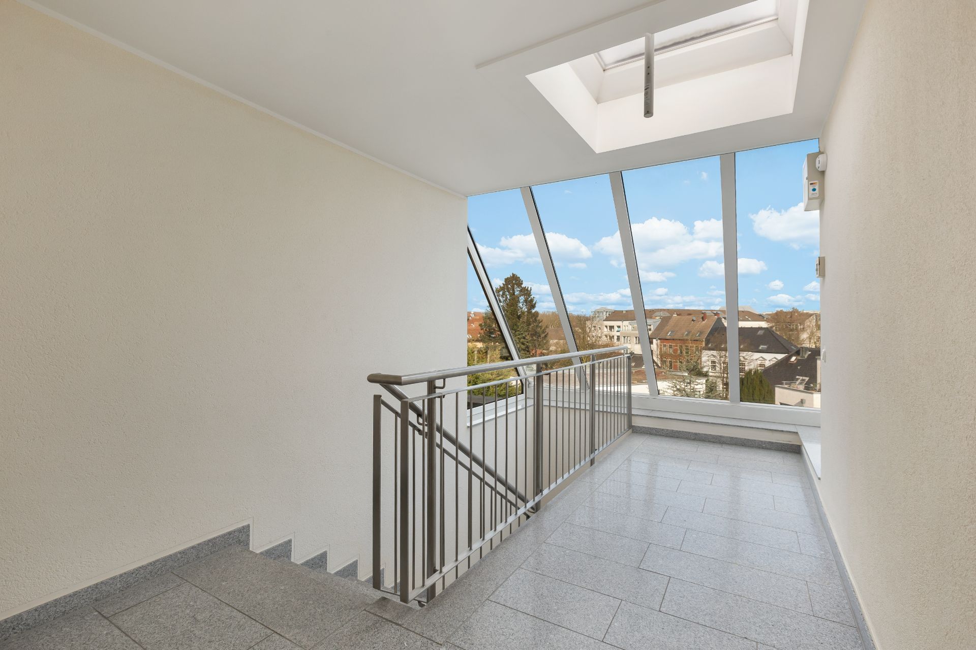 Helles Treppenhaus mit großen Fenstern und Blick auf Dächer einer Wohnsiedlung.
