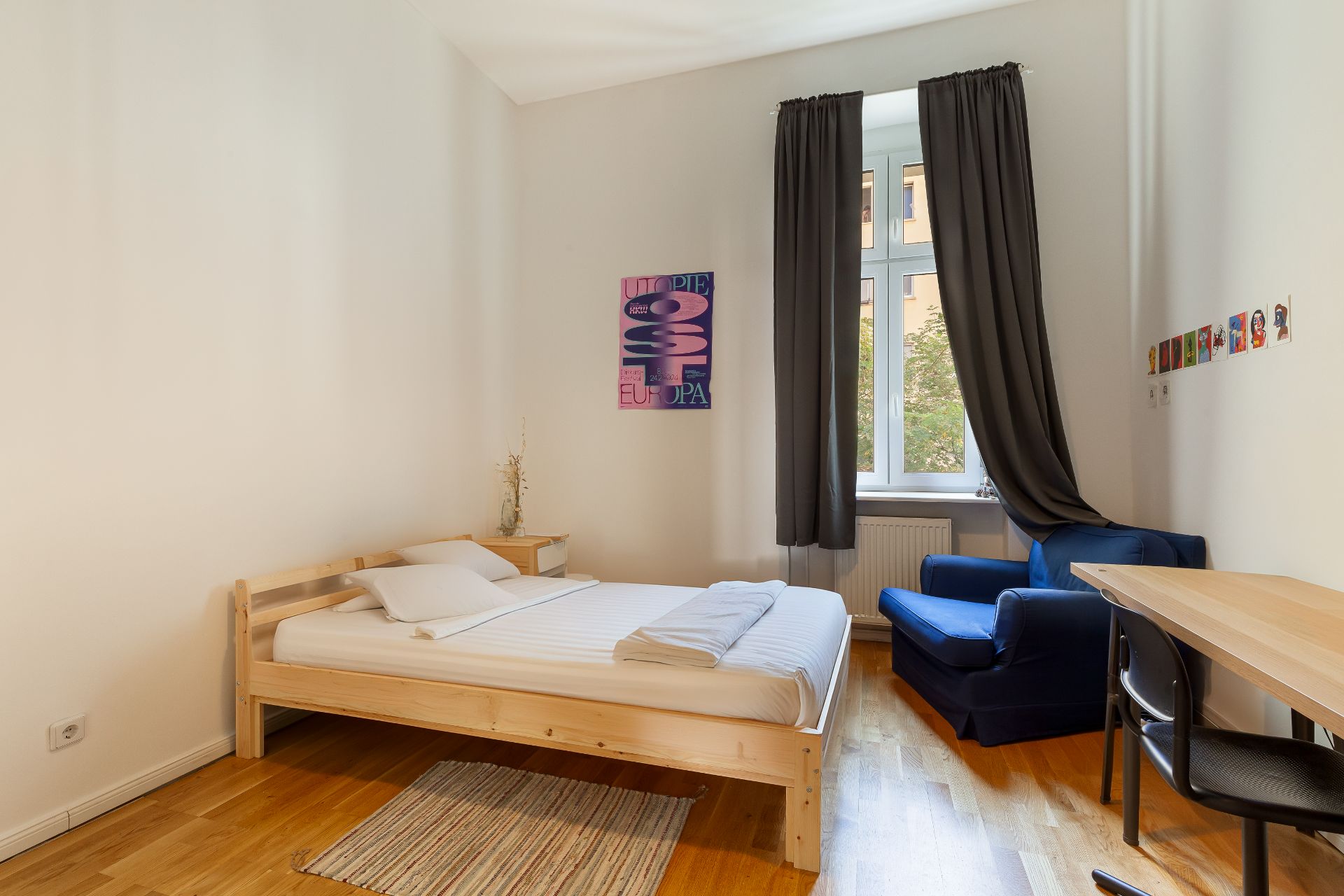 Helles Schlafzimmer mit Holzbett, Teppich, blauem Sessel, Schreibtisch und Fenster mit dunklen Vorhängen.