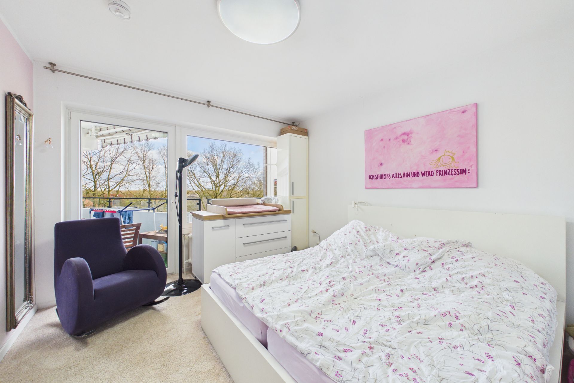 Helles Schlafzimmer mit Doppelbett, Sessel, Kommode und Zugang zu einem Balkon.