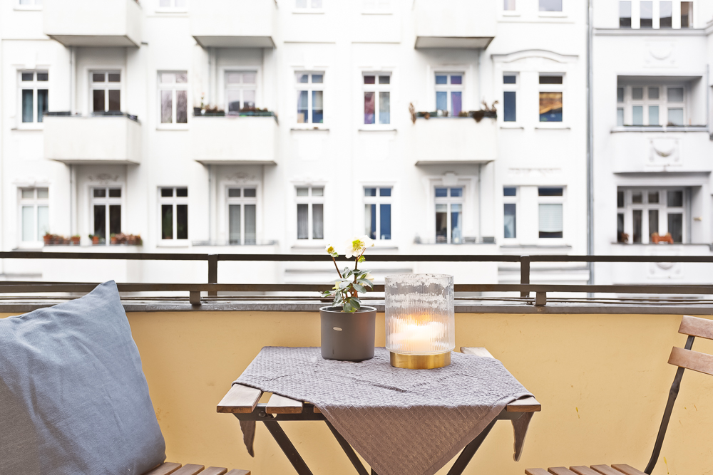 Balkon mit kleinem Tisch, Blume, Kerze und Stühlen, im Hintergrund weiße Wohnhäuser.