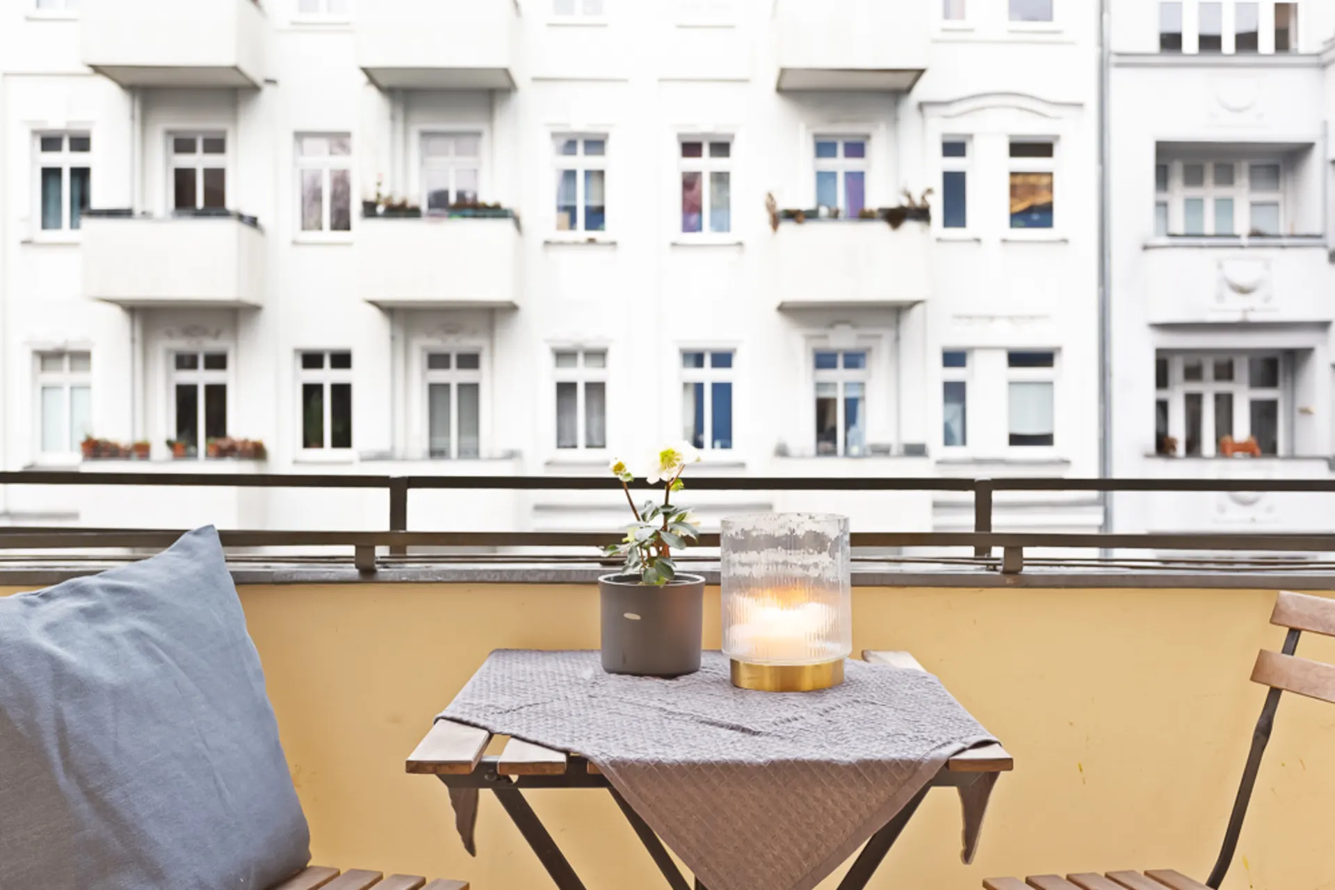 Balkon mit kleinem Tisch, Blume, Kerze und Stühlen, im Hintergrund weiße Wohnhäuser.