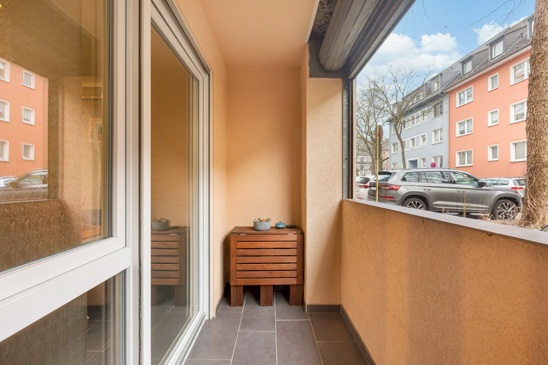 Balkon mit Holzbank und kleiner Pflanze, Blick auf Straße und Wohnhäuser.