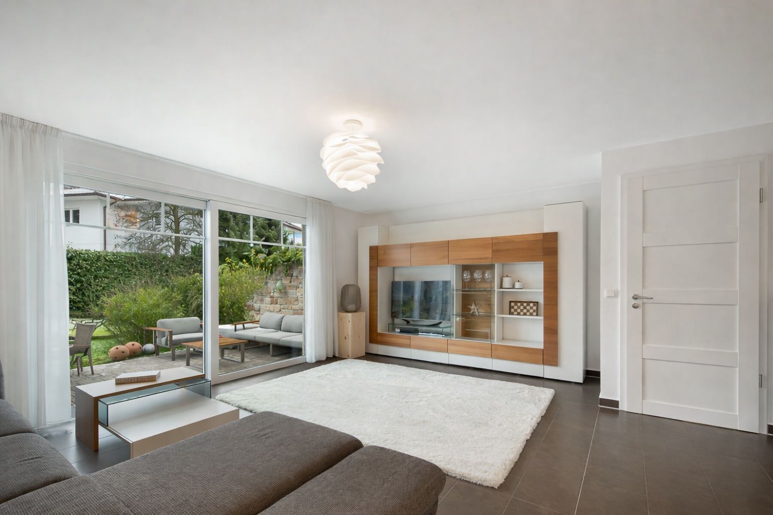 Helles Wohnzimmer mit großem Fenster, grauem Sofa, TV-Wand und Teppich, Blick auf Terrasse und Garten.