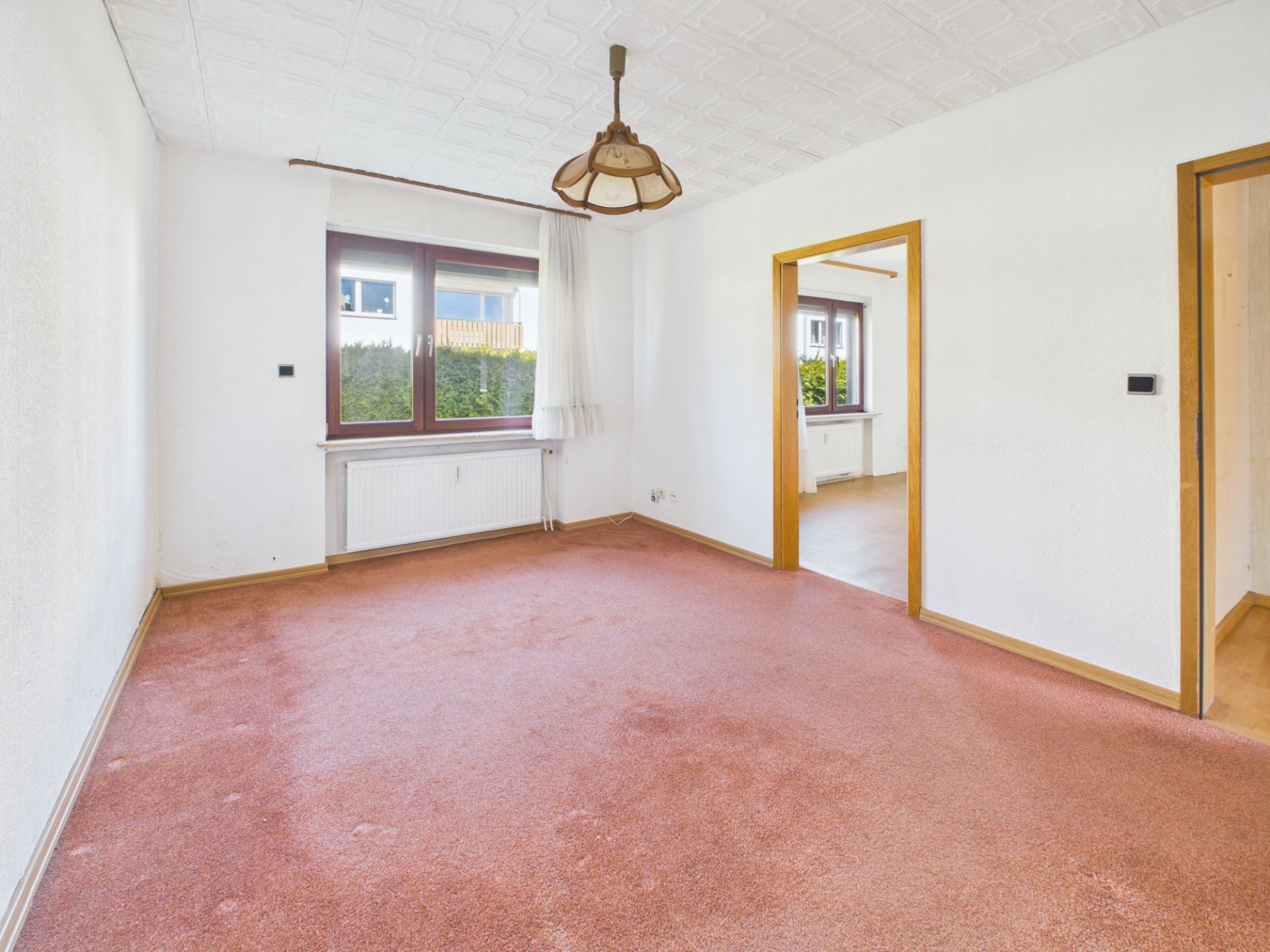 Leeres Wohnzimmer mit rotem Teppich, weißer Decke, Fenster und zwei offenen Türen.