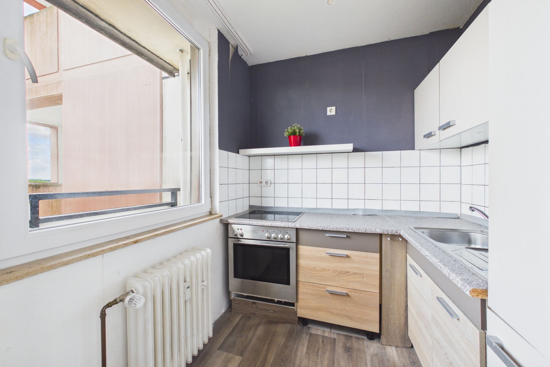 Helle Küche mit Fenster, Herd, Spüle, weißen Fliesen und kleinem Pflanztopf auf dem Regal.