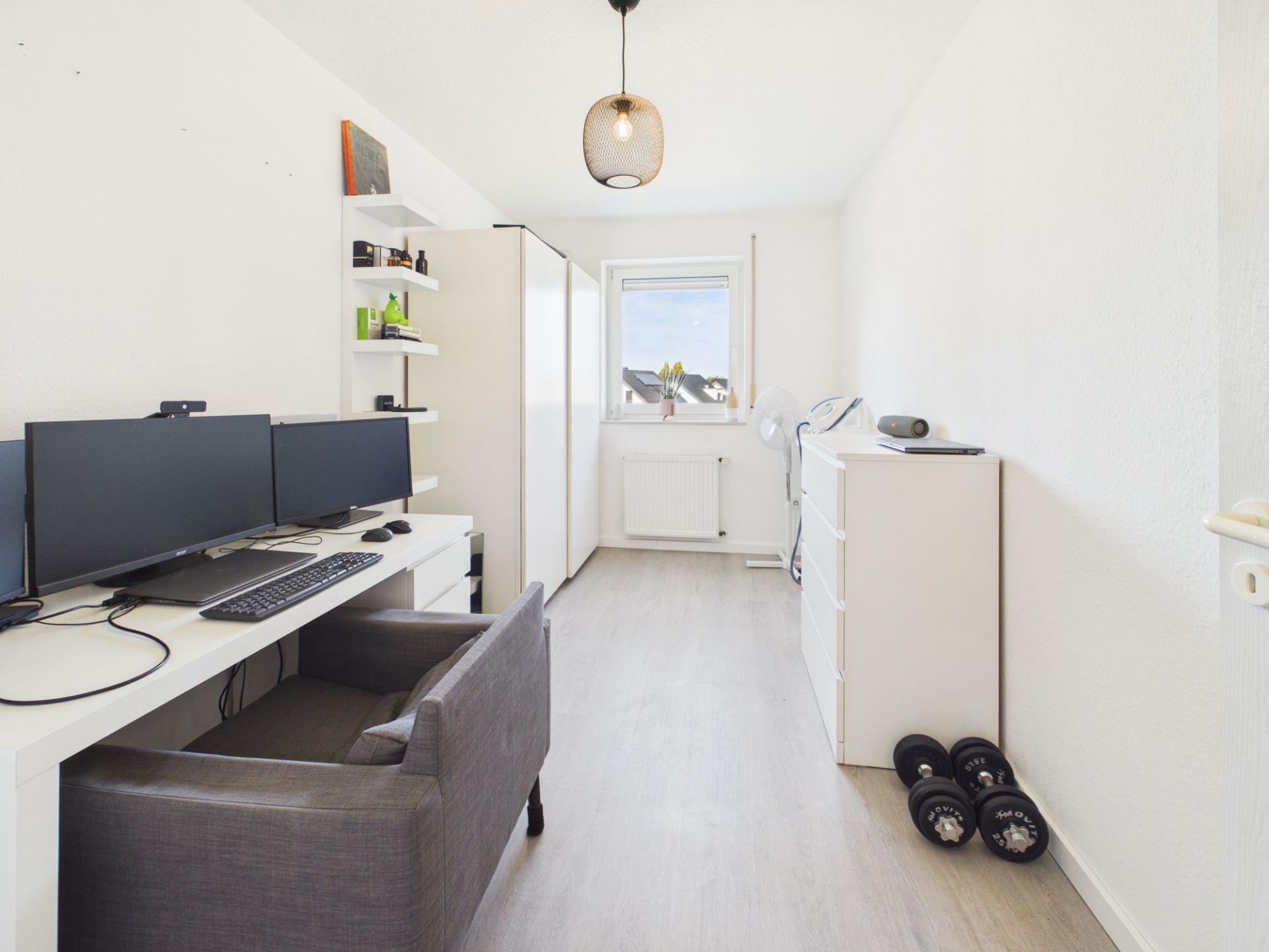 Helles Arbeitszimmer mit Schreibtisch, Monitoren, Regal, Kommode und Hanteln auf hellem Boden.