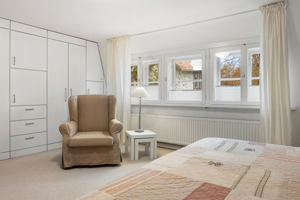 Helles Schlafzimmer mit Sessel, weißem Schrank, Beistelltisch und drei Fenstern mit Vorhängen.