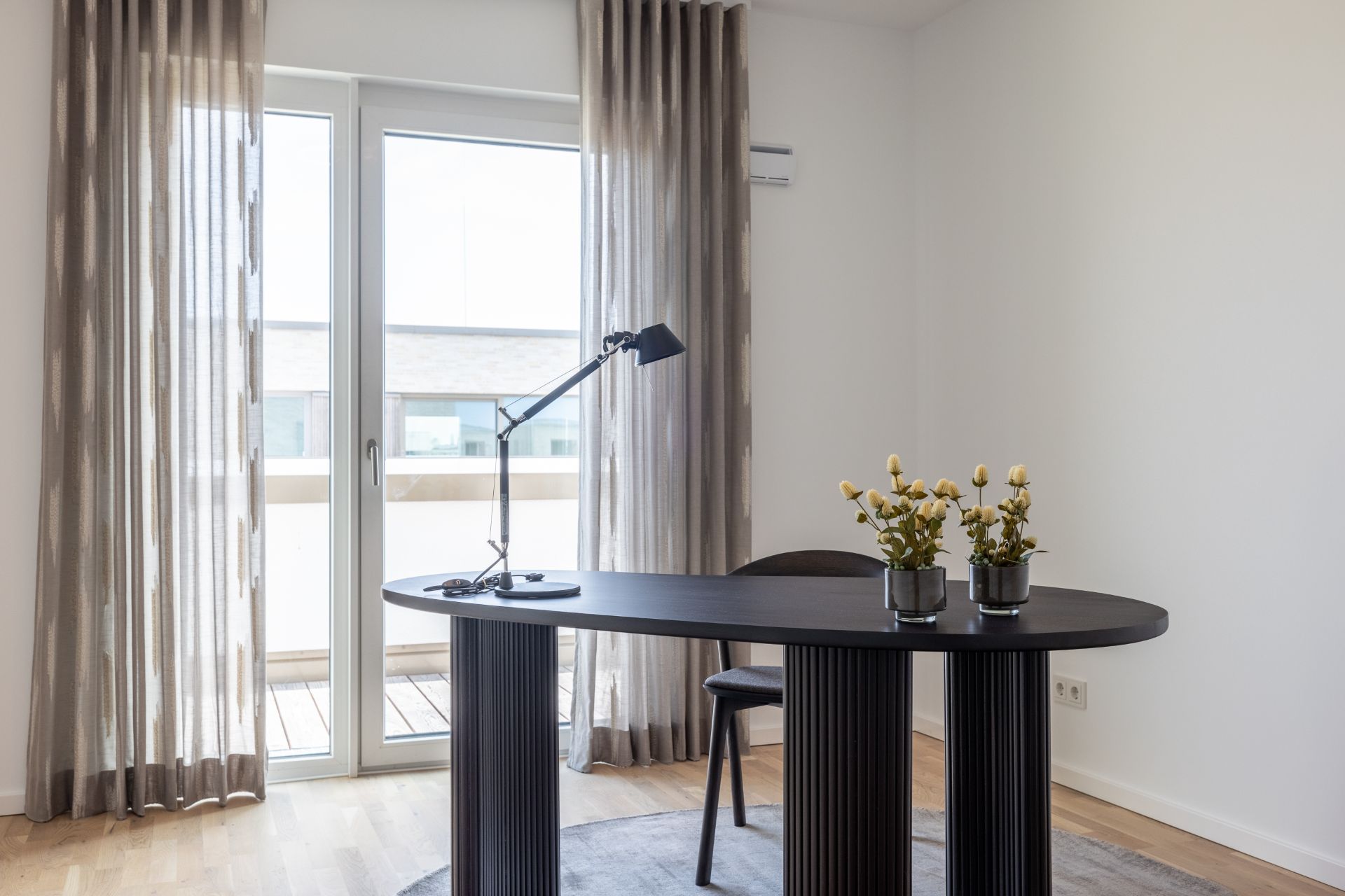 Moderner Schreibtisch mit Lampe und Pflanzen vor bodentiefen Fenstern mit Vorhängen.