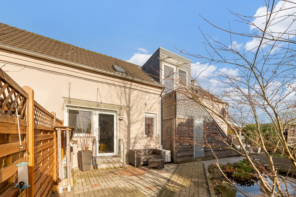 Hauseingang mit Terrasse, Sitzgelegenheit, Holzzaun und kleinem Teich im Vordergrund.