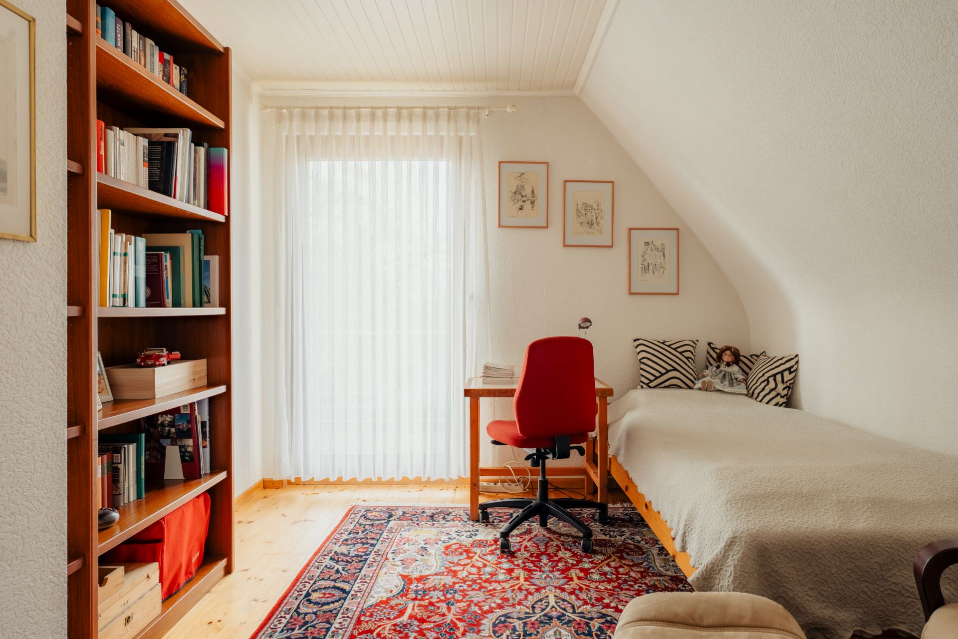Helles Zimmer mit Bett, Schreibtisch, rotem Stuhl, Bücherregal und Teppich.