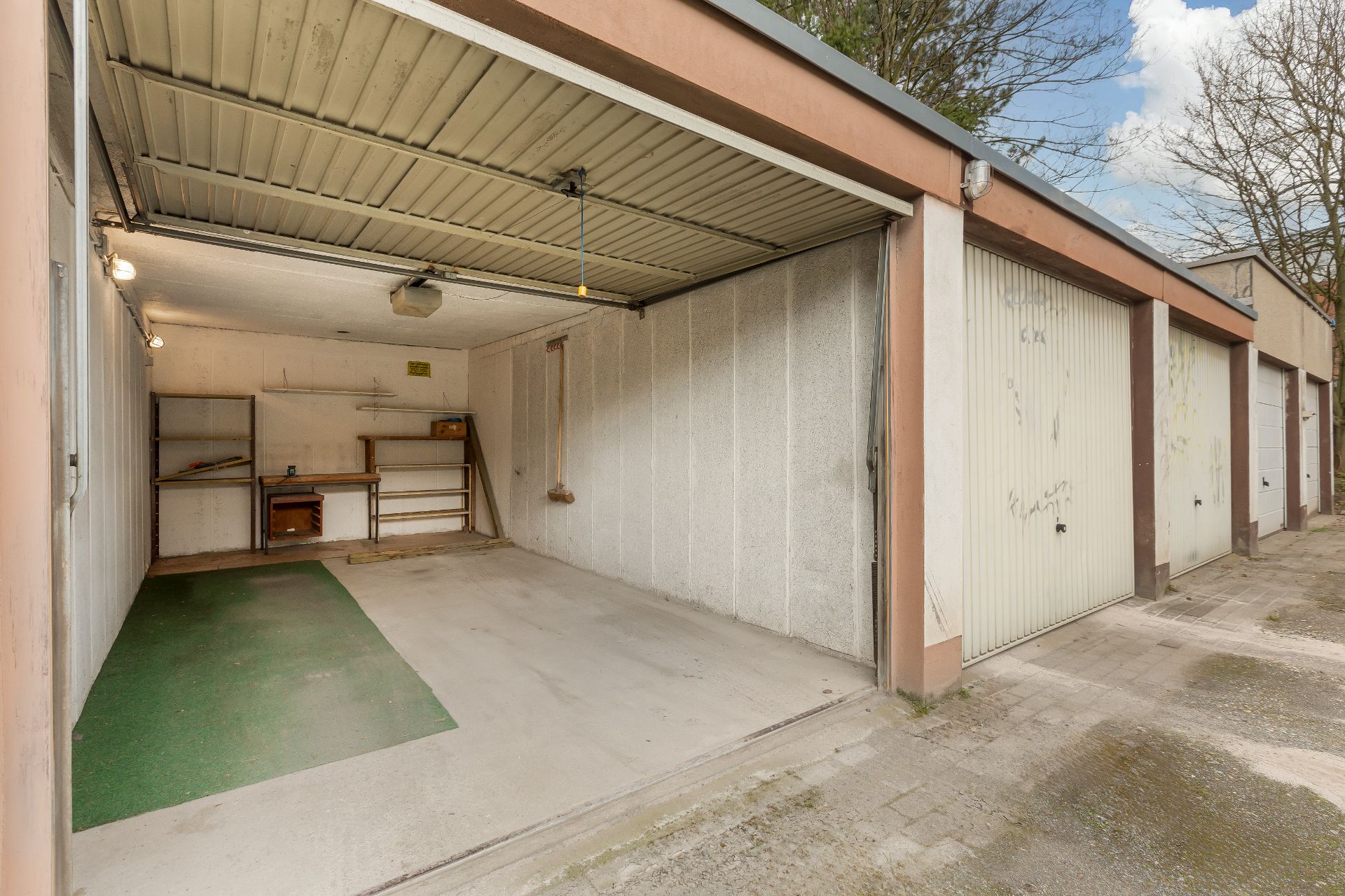 Offene Garage mit Betonboden, grünem Teppichstück und Regal an der Rückwand.
