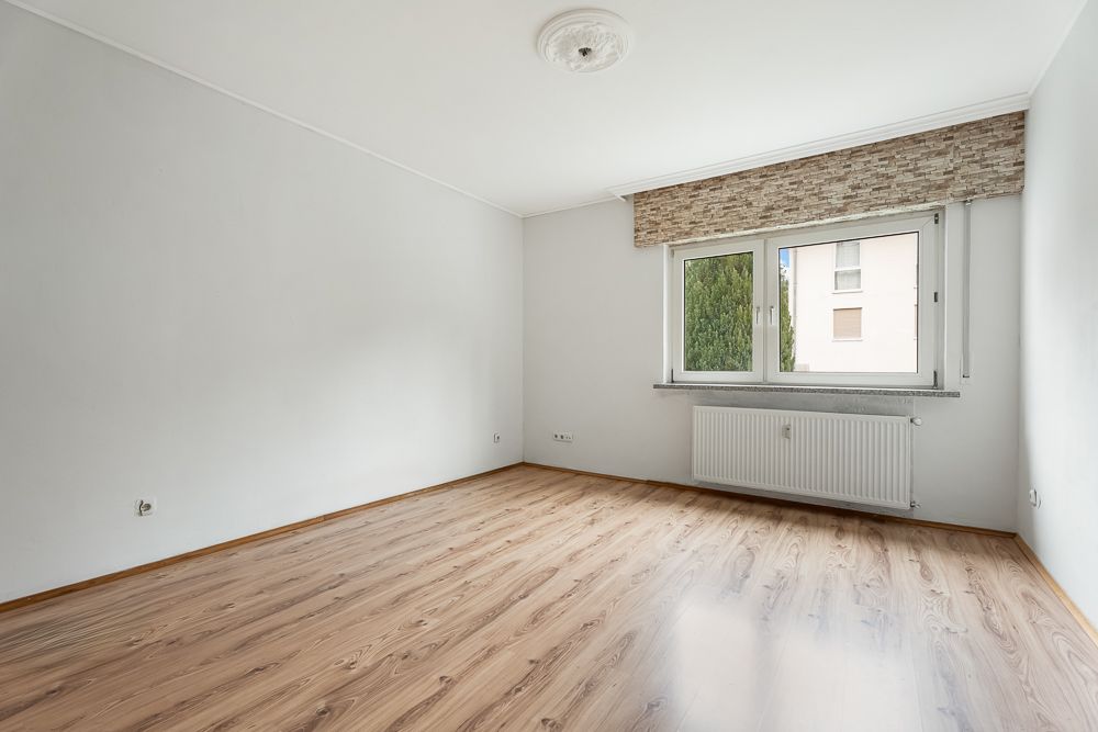 Leerer Raum mit Holzfußboden, weiß gestrichenen Wänden und großem Fenster mit Heizung darunter.