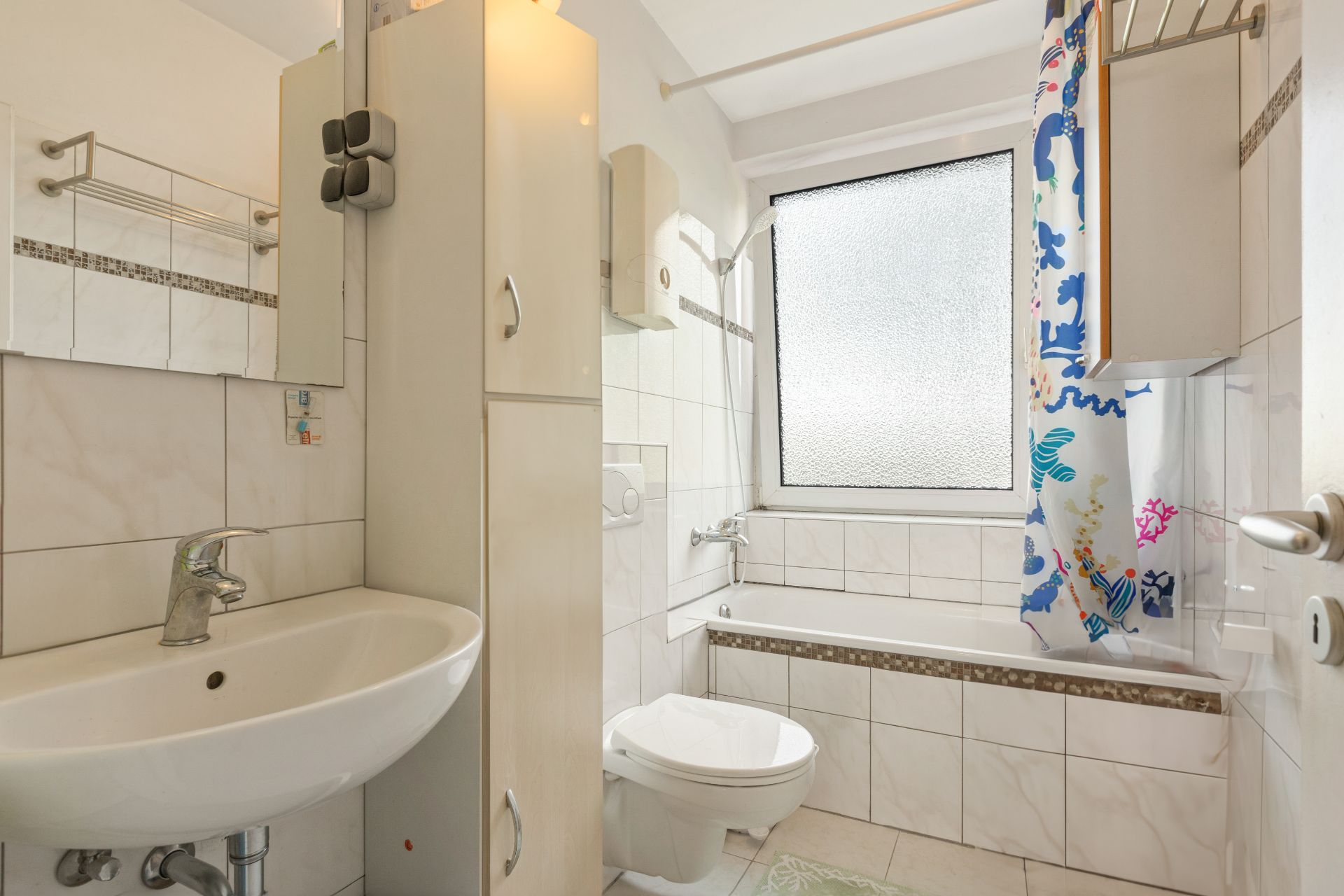 Helles Bad mit Waschbecken, WC, Badewanne und Duschvorhang mit buntem Muster.