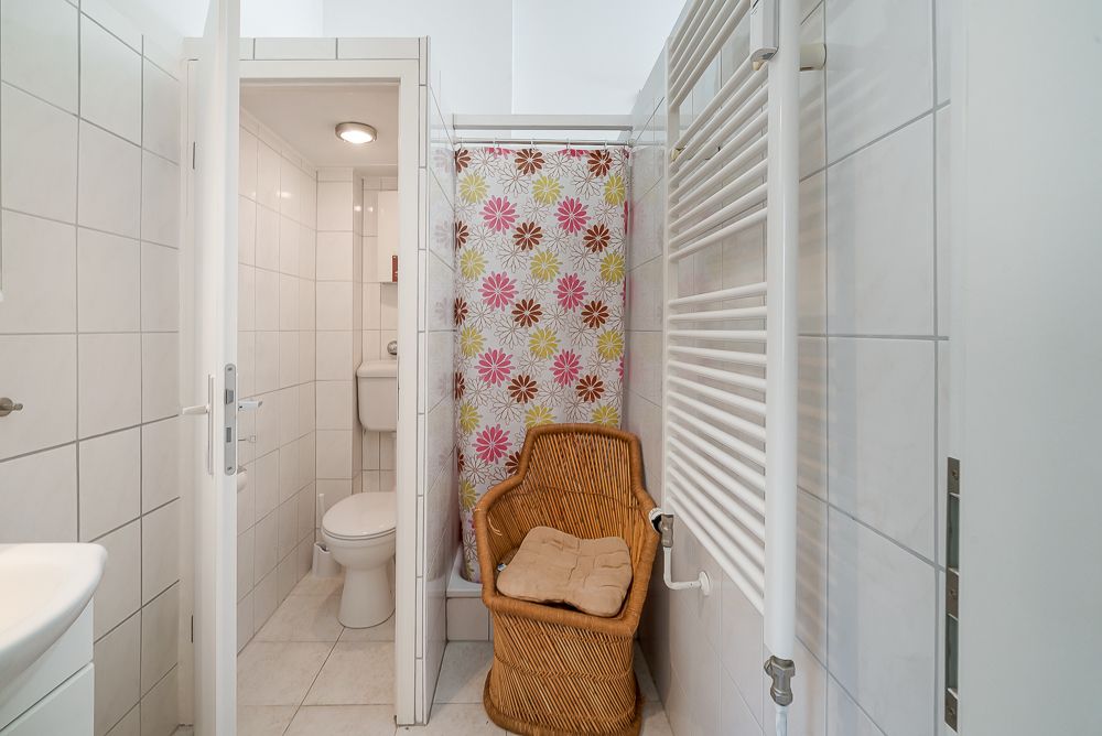 Kleines Badezimmer mit Dusche, bunter Duschvorhang, Toilette und Korbstuhl.