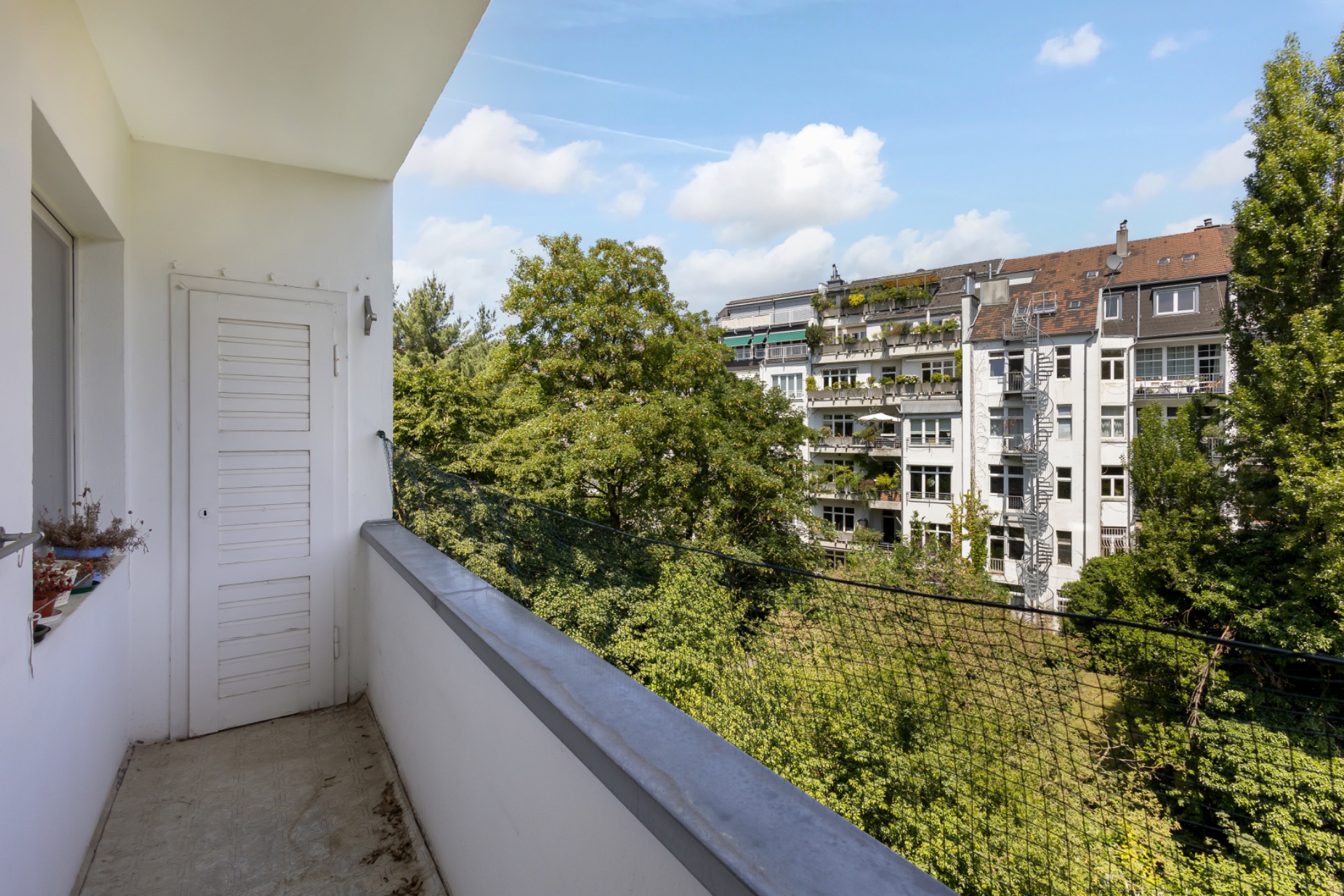 Balkon mit Blick auf Bäume und gegenüberliegende Wohngebäude an einem sonnigen Tag.
