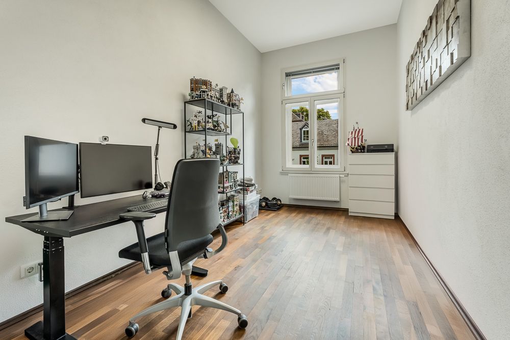 Helles Arbeitszimmer mit Schreibtisch, zwei Monitoren, Regal und Fenster, Holzboden.