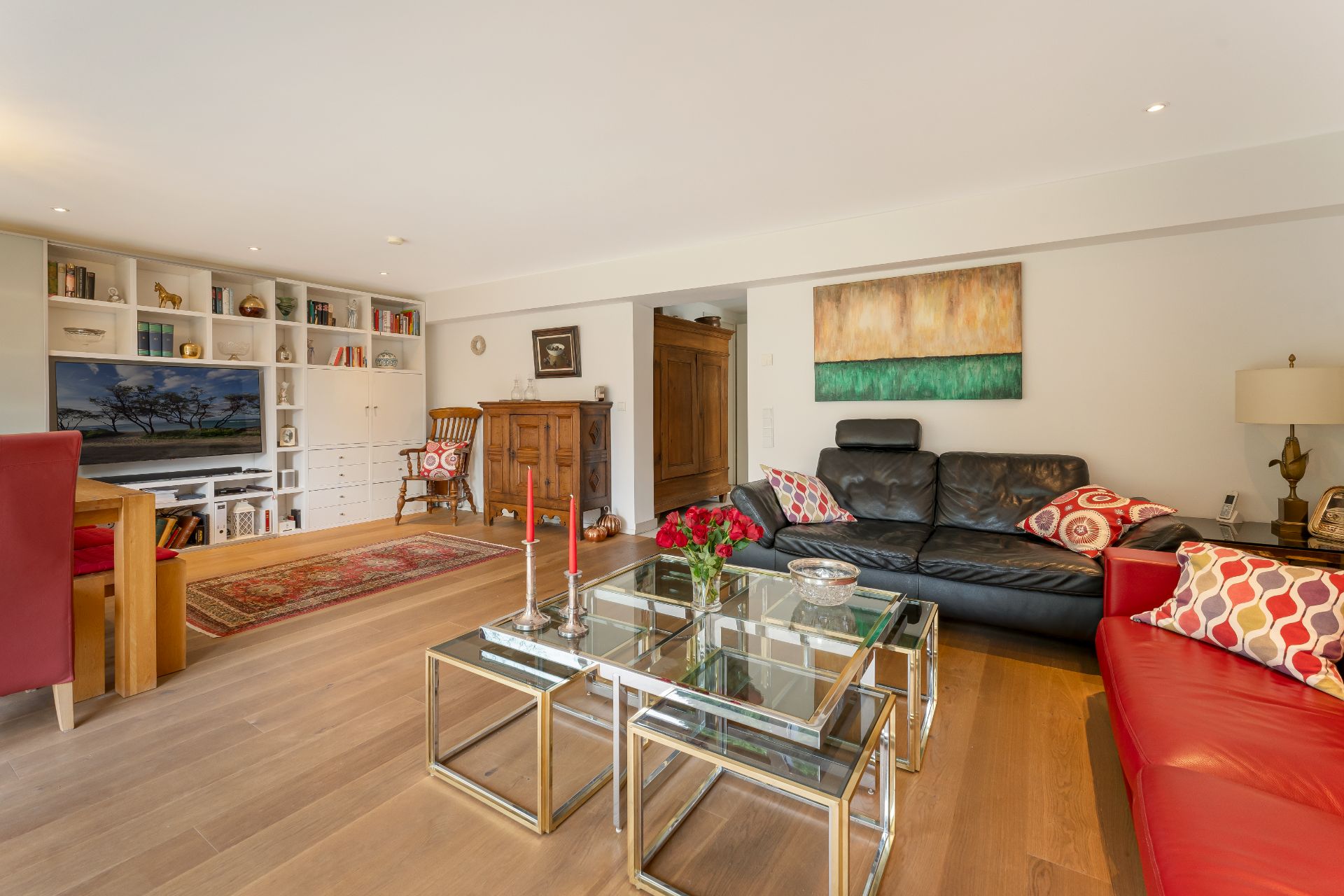 Helles Wohnzimmer mit schwarzem Sofa, rotem Sofa, Glastisch und Regal mit Fernseher.