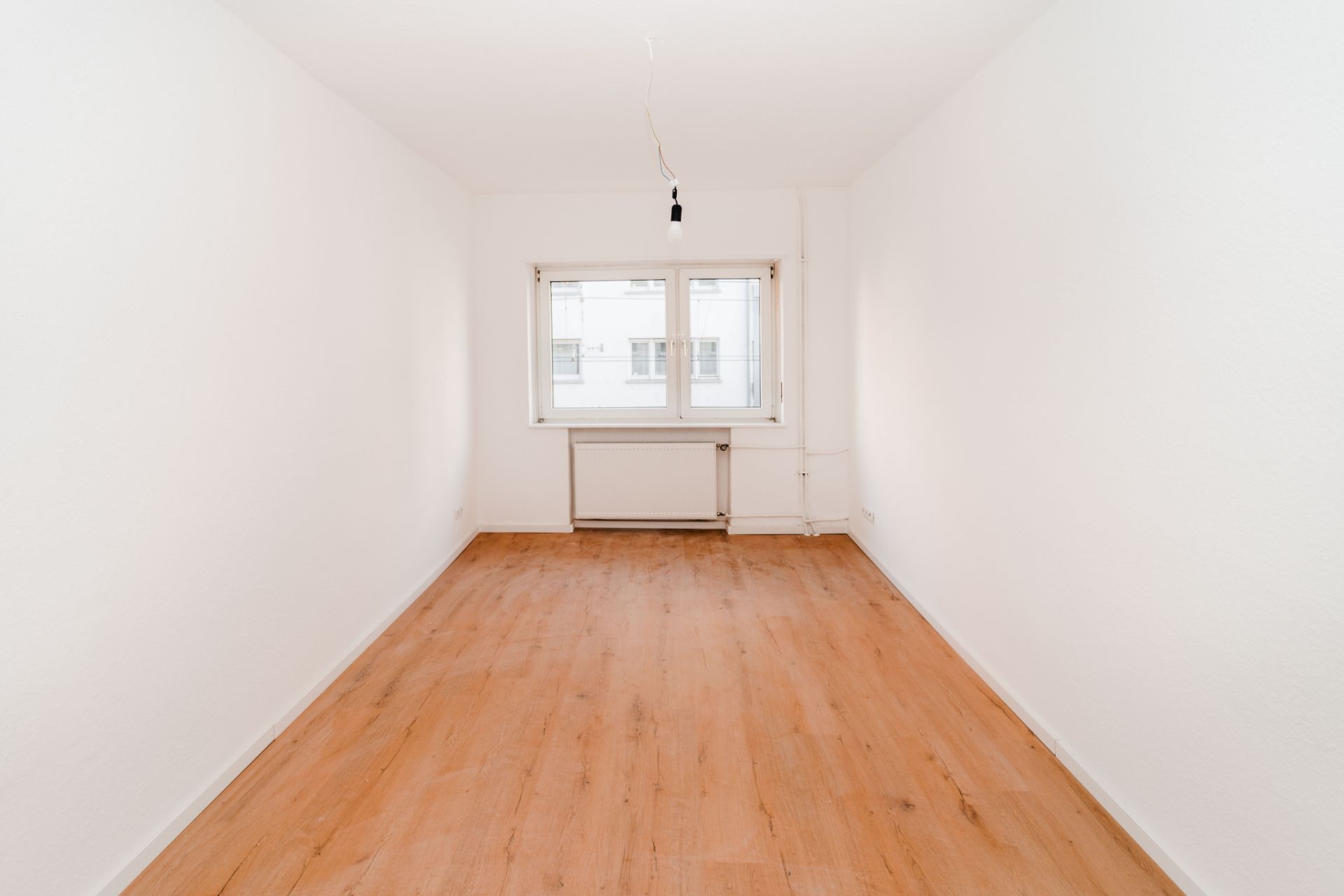 Leerer Raum mit Holzfußboden, weiß gestrichenen Wänden und einem Fenster am Ende.