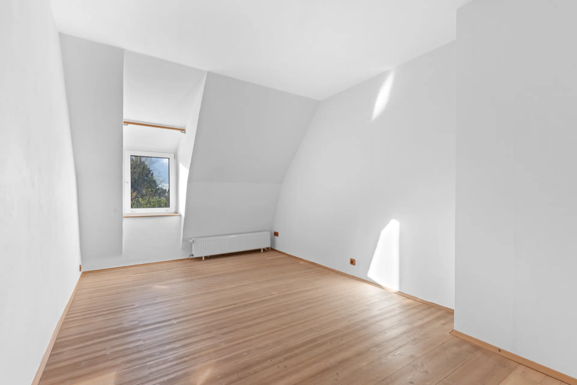 Helles, leeres Zimmer mit Holzfußboden, weißer Wand und einem Fenster mit Blick ins Grüne.