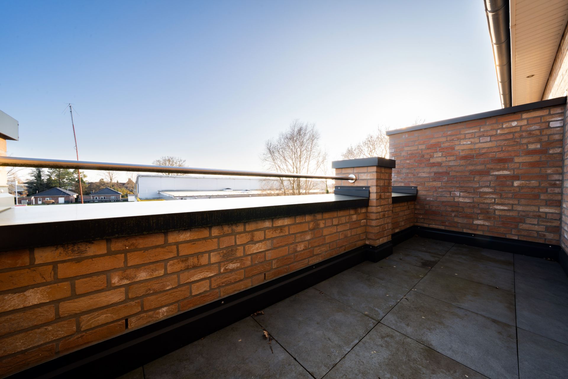 Große Terrasse mit Backsteinmauer und Metallgeländer, Blick auf Bäume und blauen Himmel.