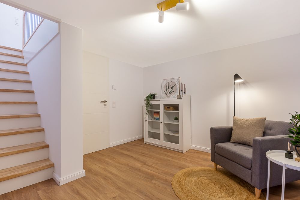 Helles Wohnzimmer mit grauem Sessel, Teppich, Regal, Lampe und Holztreppe.