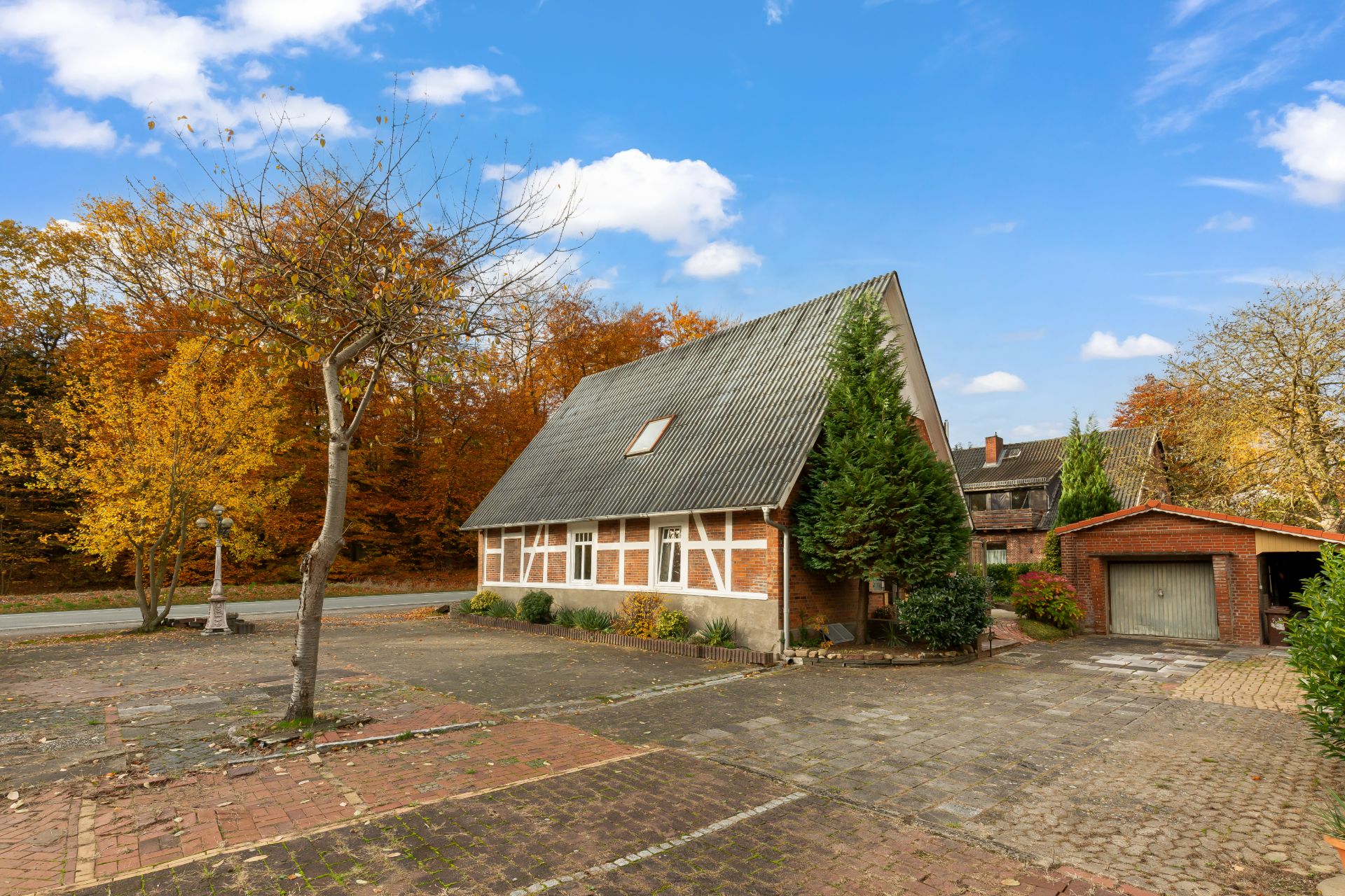 Fachwerkhaus mit steilem Dach, Bäumen und gepflastertem Hof vor herbstlichem Wald.