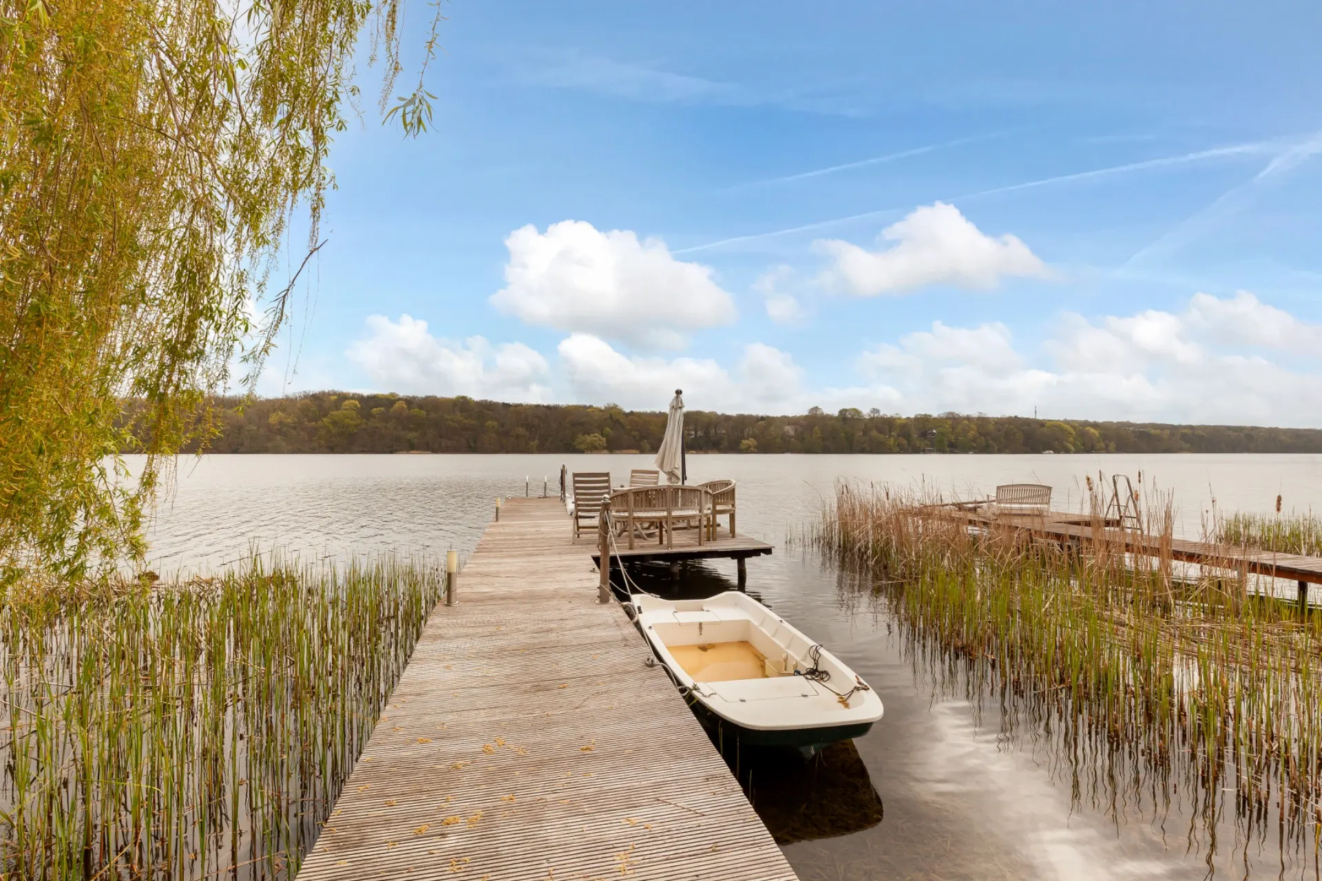 Steg mit kleinem Boot und Sitzbereich am Ufer eines Sees, umgeben von Schilf und Bäumen.