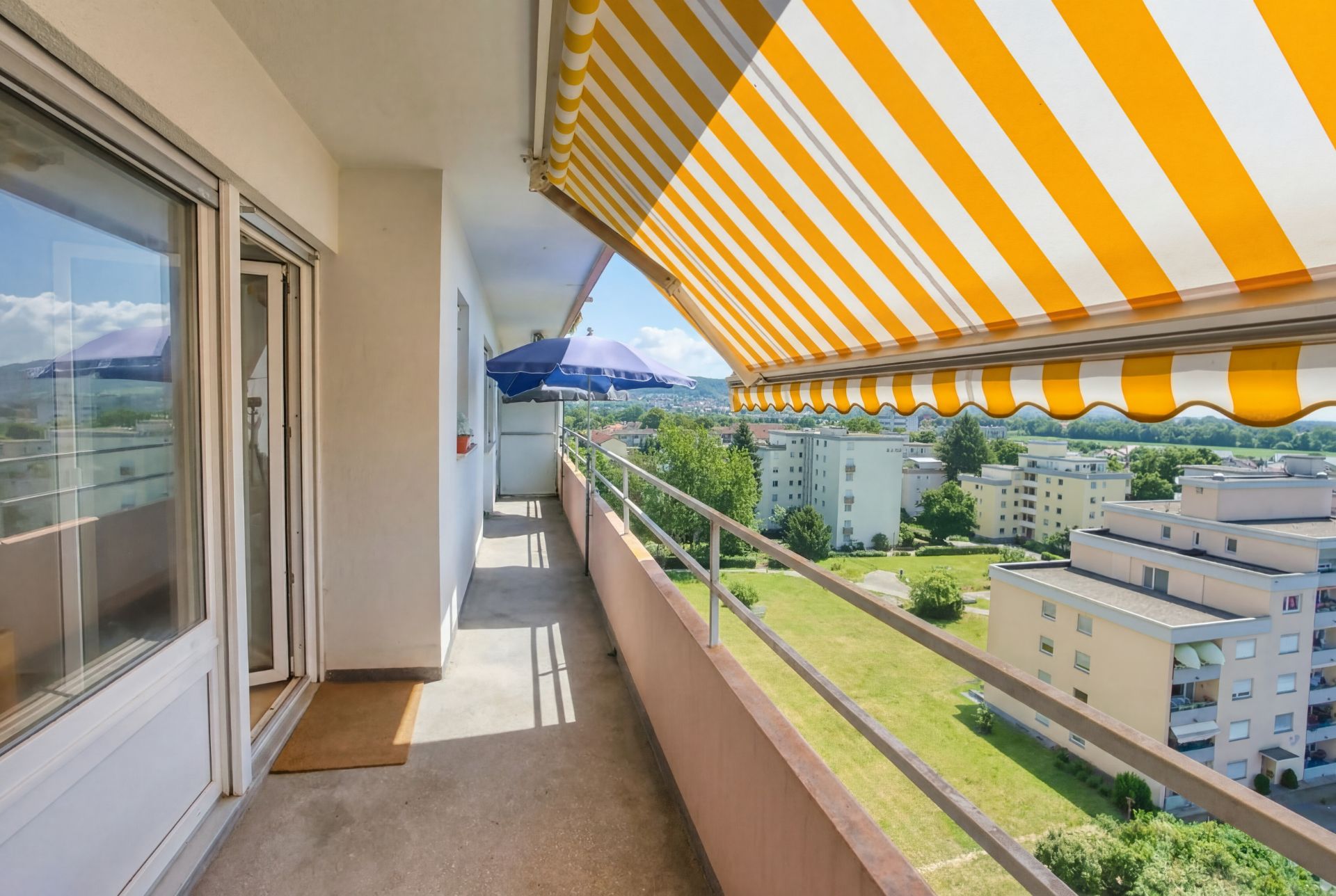 Langer Balkon mit gelb-weißer Markise und Blick auf Wohnhäuser und Grünflächen.