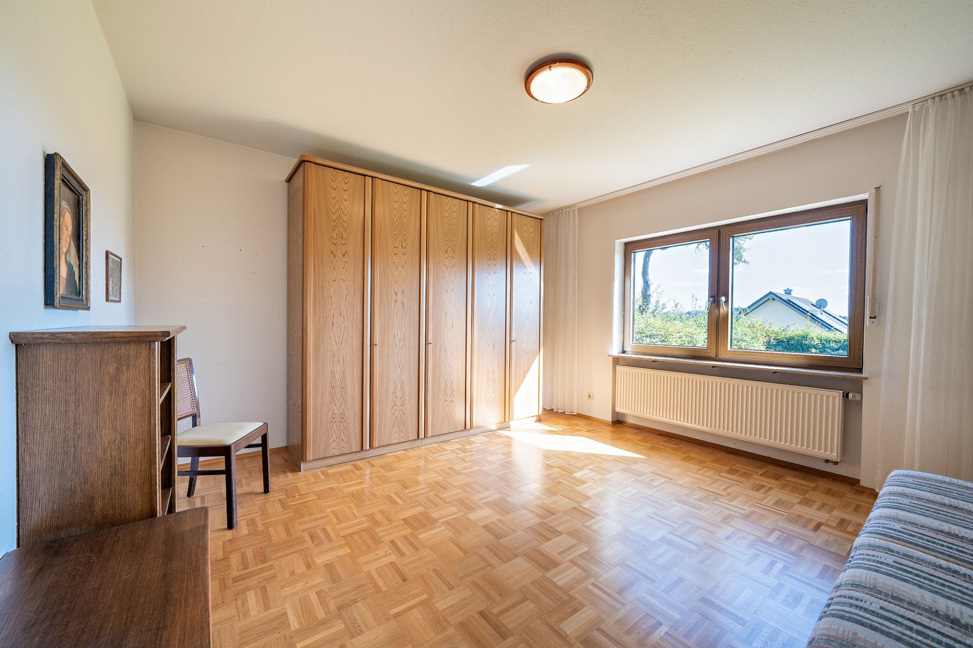 Helles Zimmer mit Holzschrank, Fenster, Parkettboden, Kommode und Stuhl.