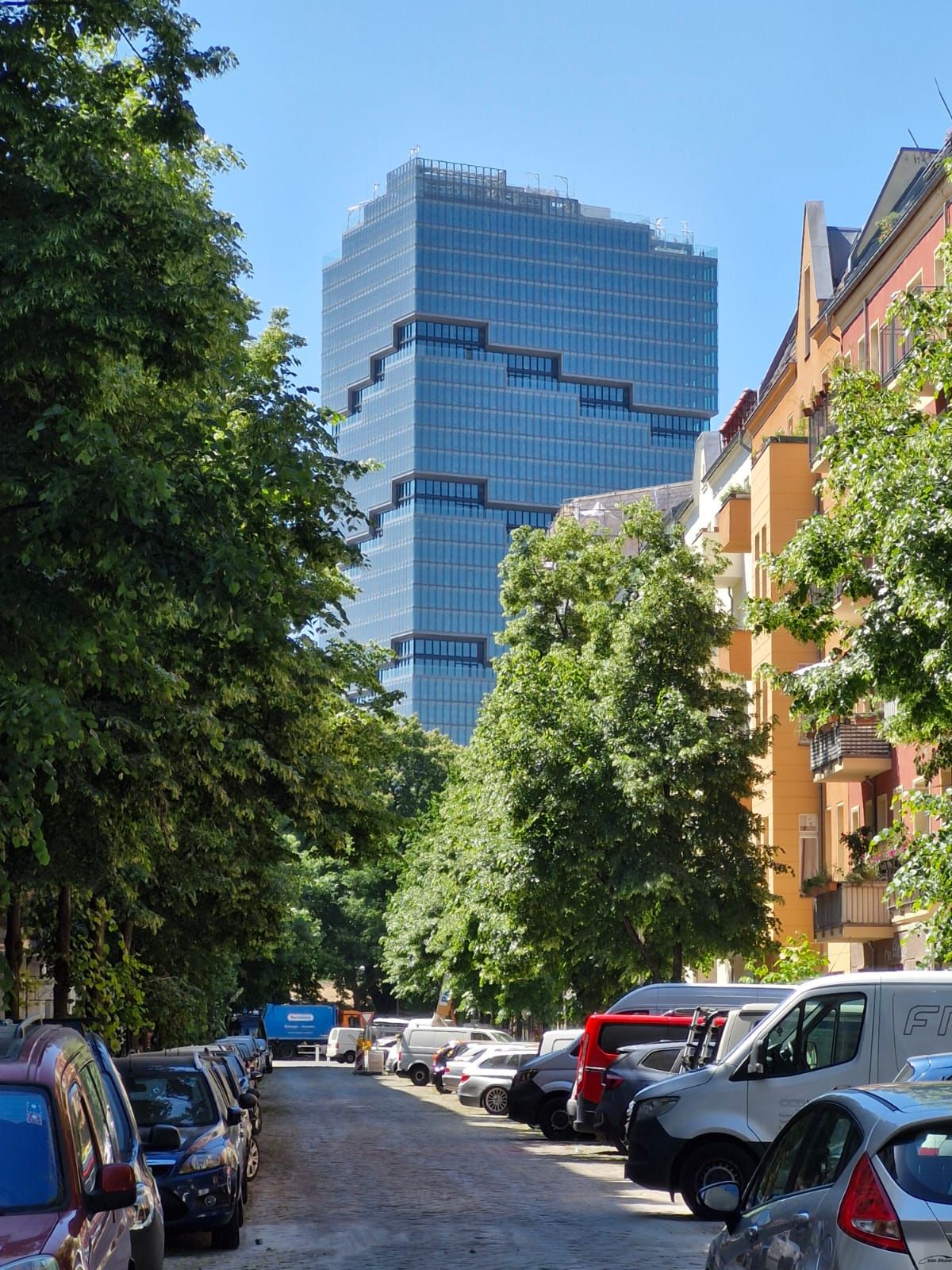Wohnstraße mit parkenden Autos, Bäumen und modernem Glasgebäude im Hintergrund.