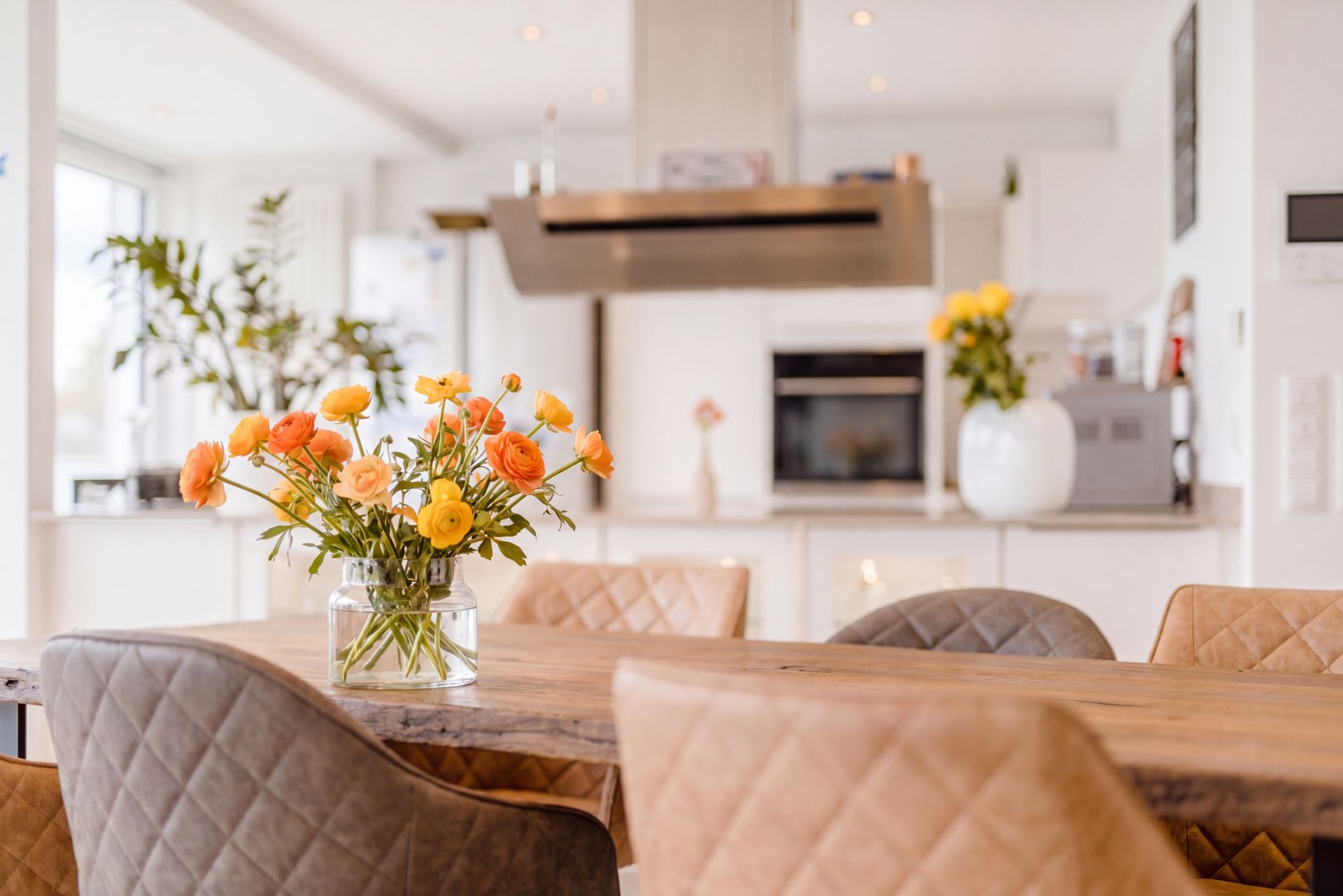 Essbereich mit Holztisch, gepolsterten Stühlen und Vase mit bunten Blumen, dahinter moderne Küche.