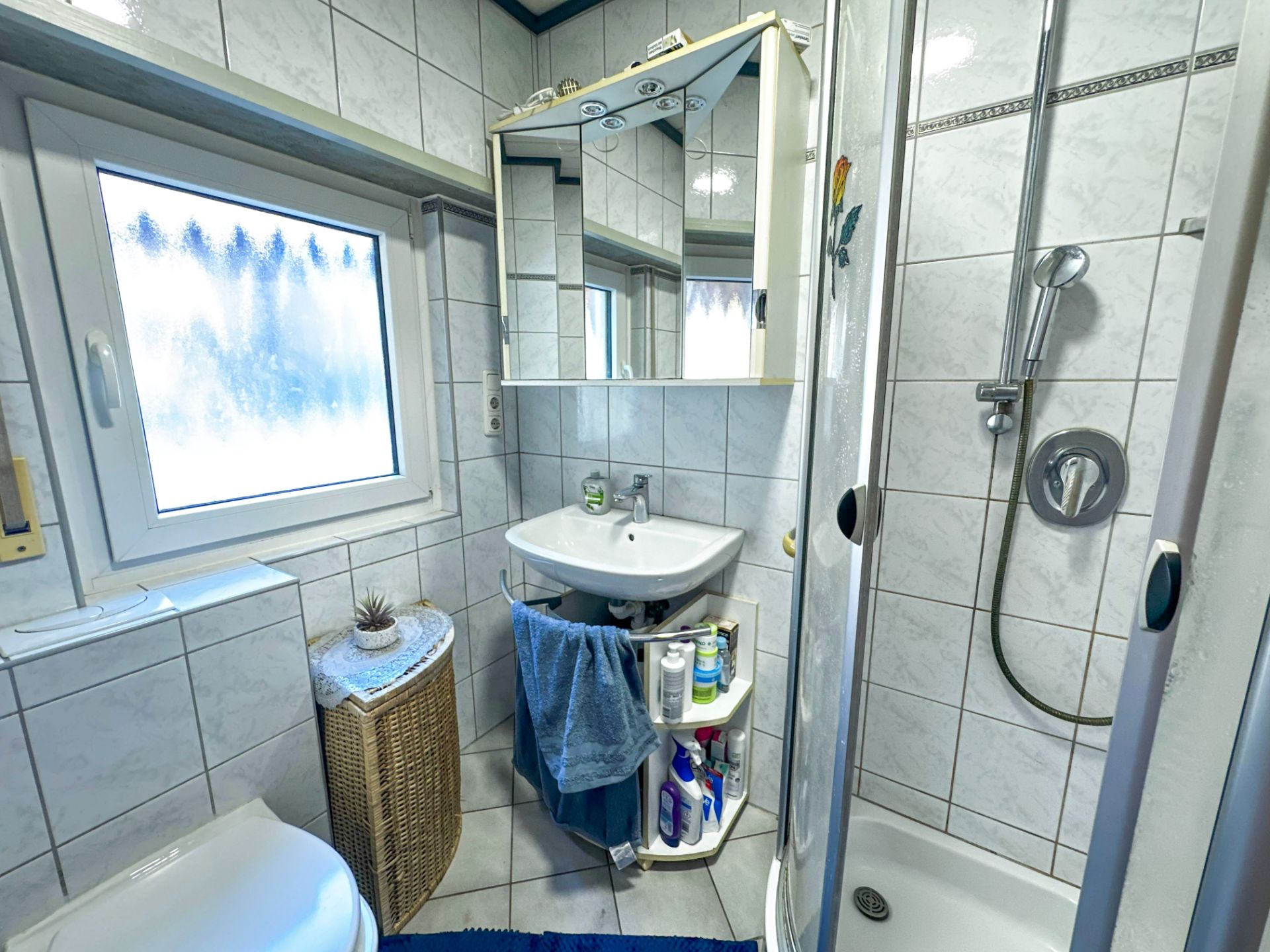 Kleines, gefliestes Bad mit Dusche, Waschbecken, WC, Spiegel und Fenster.