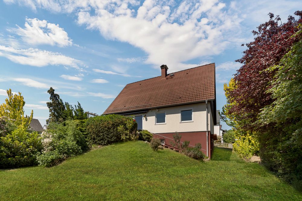 Ein Einfamilienhaus mit Garten, Rasenfläche und umliegenden Büschen unter blauem Himmel.